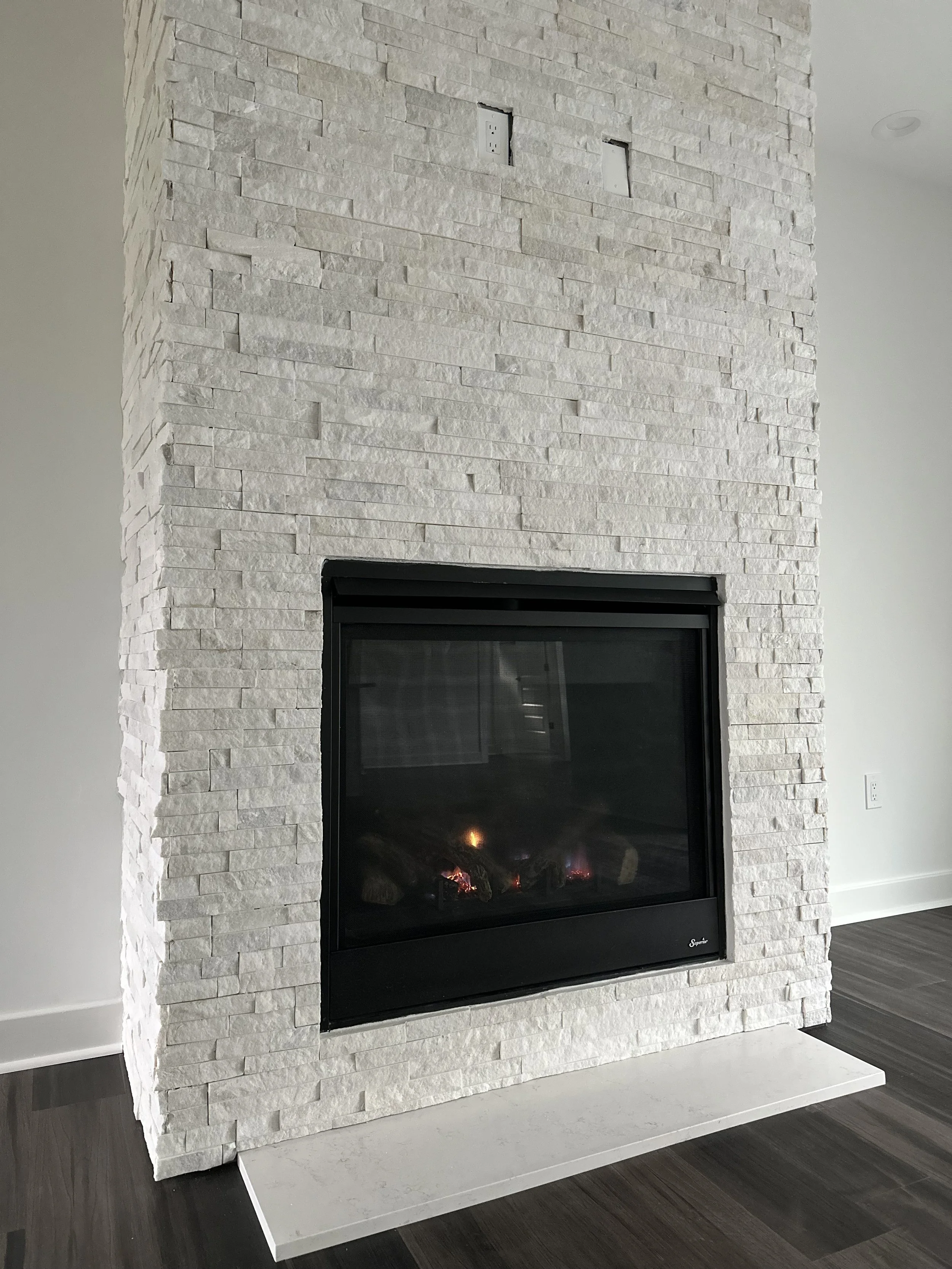 White stone fireplace with a black glass front, located on a dark wood floor, with empty electrical outlets above.