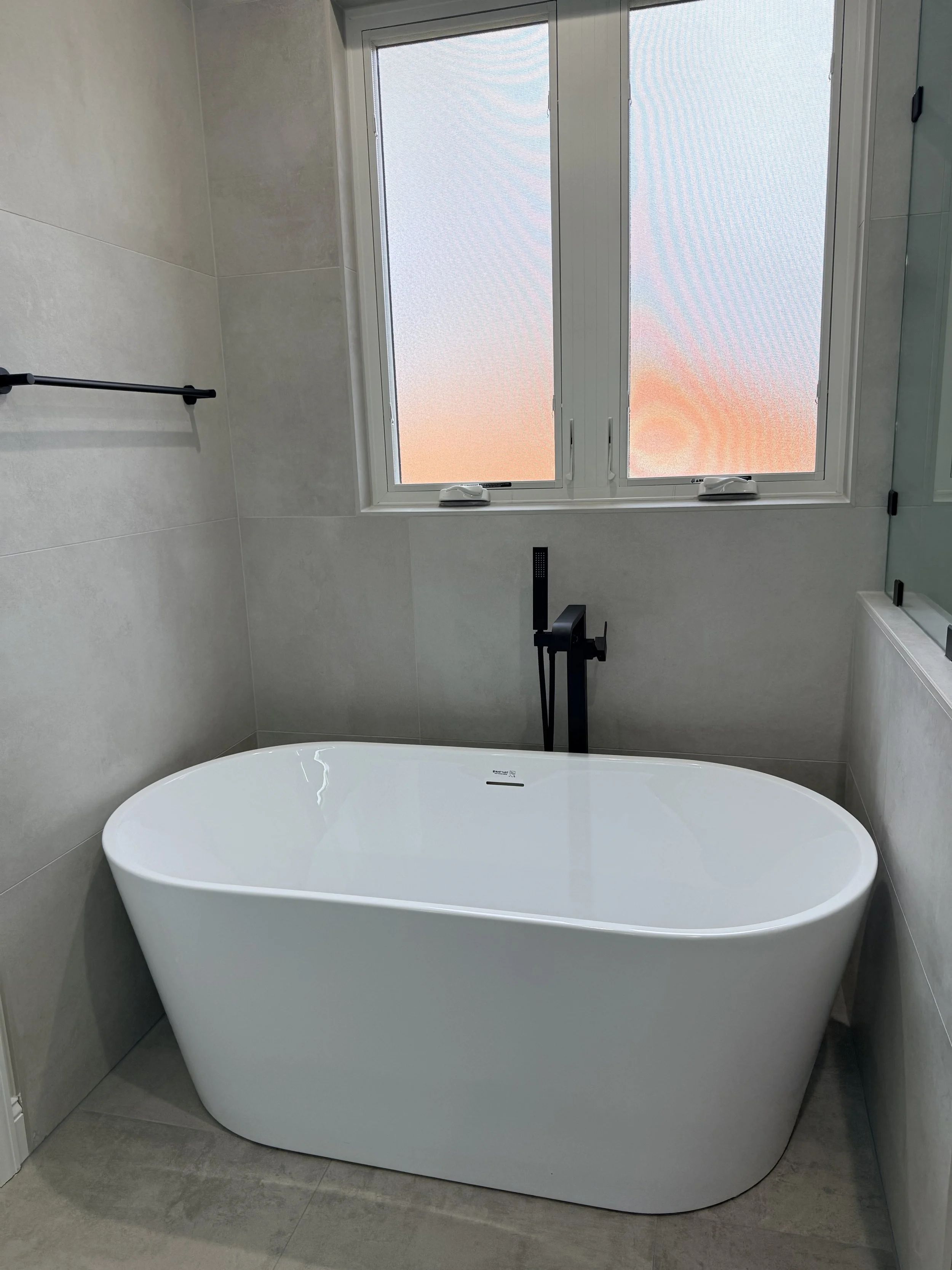 Modern bathroom with a white freestanding bathtub, gray tiled walls, a large window showing a sunset sky, and a black faucet.