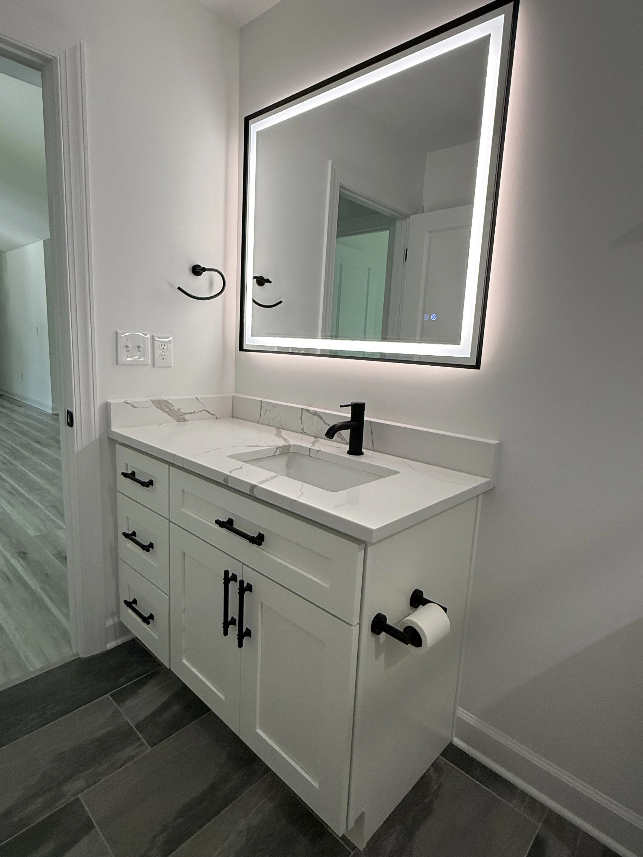 Modern bathroom vanity with white cabinet, marble countertop, black faucet, large backlit mirror, and toilet paper holder on the side.