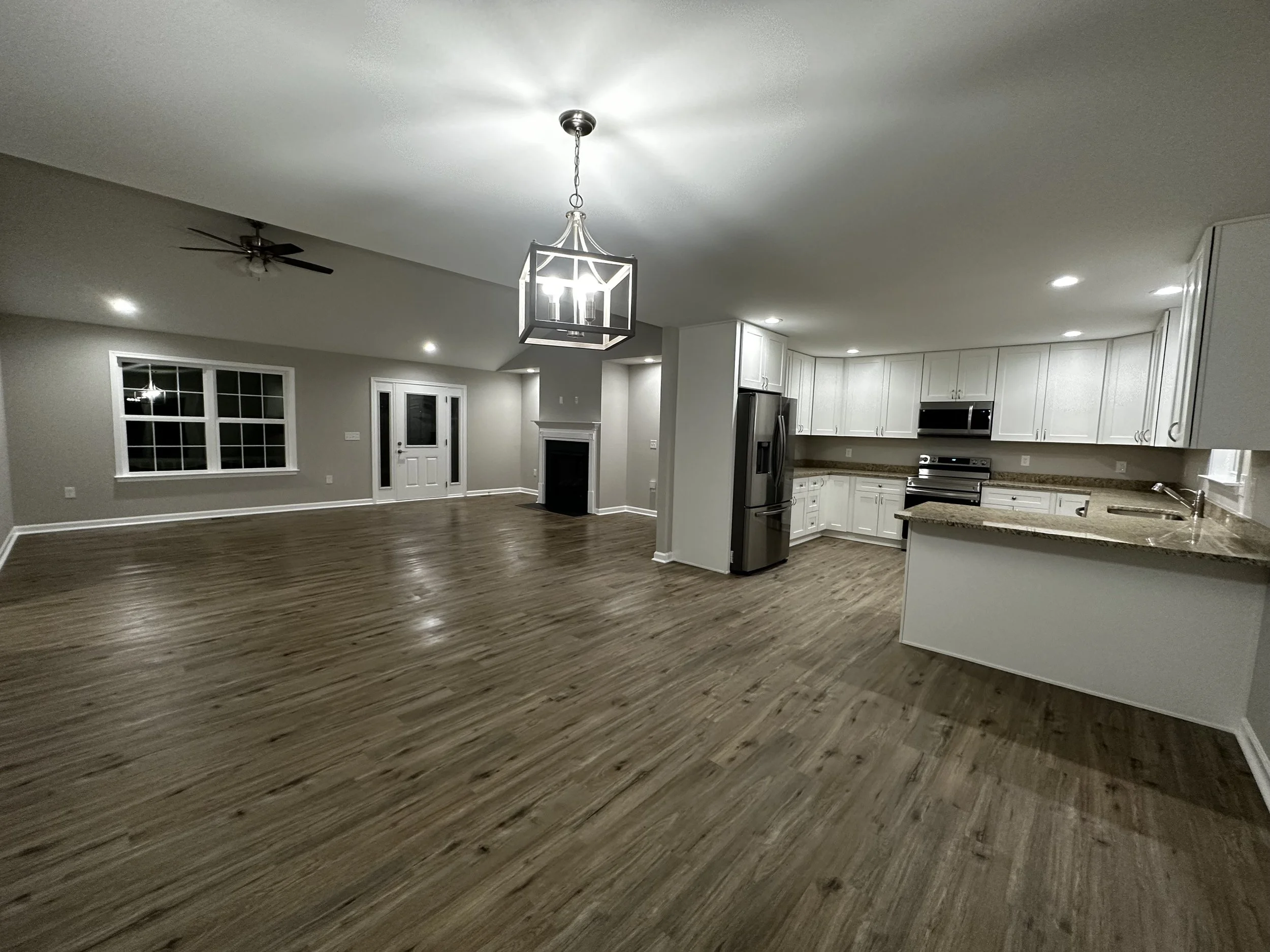 Empty open-concept living room and kitchen with hardwood floors, white cabinets, stainless steel appliances, a fireplace, large window, and modern lighting fixtures.