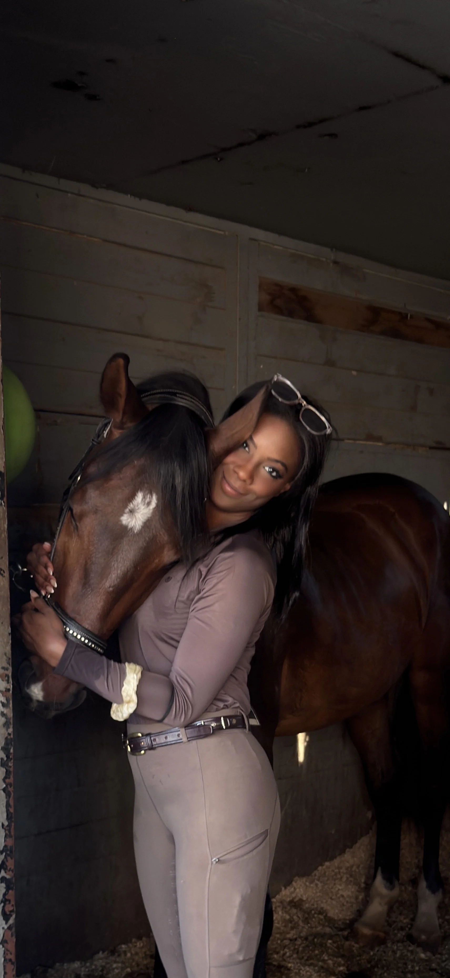 A woman hugging a brown horse inside a stable, with another horse partially visible on the right.