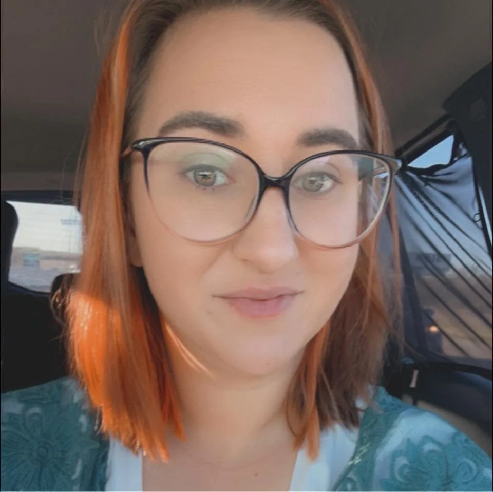 A woman with red hair and glasses taking a selfie in a car, with a view of the outside through the window.