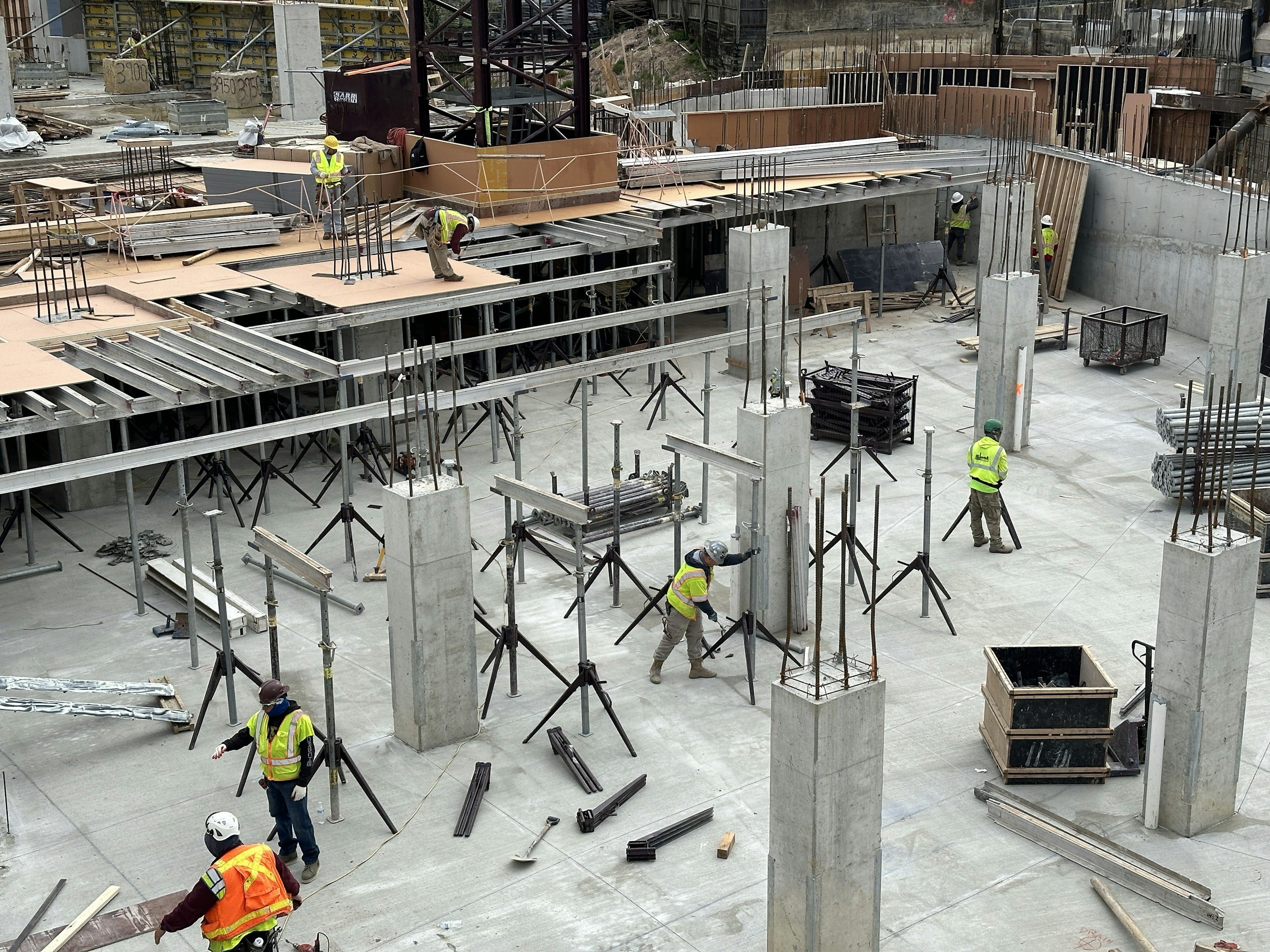 Construction workers wearing safety vests and helmets working on a building site with concrete pillars, steel rods, and various construction materials.