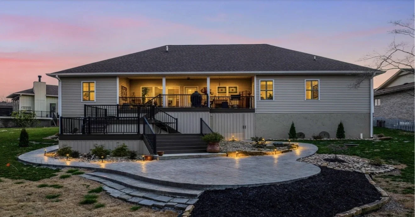 A house with a large backyard, a paved patio area, and outdoor lighting at sunset.