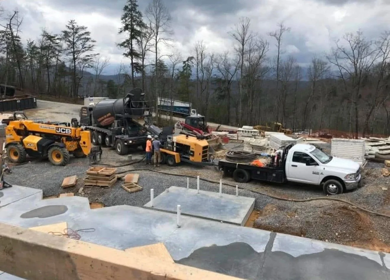  Knoxville Concrete Pumping Construction site with construction vehicles and workers, wood pallets, concrete foundation, and a wooded background with cloudy sky.