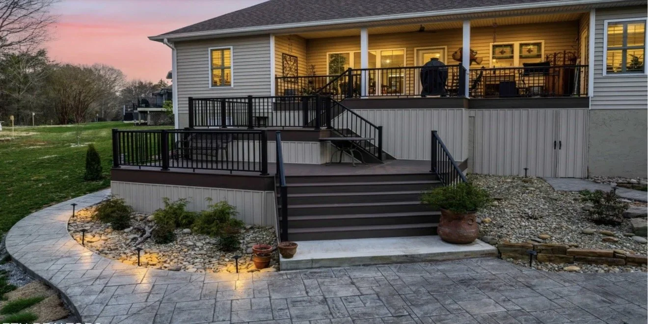 Knoxville Concrete Pumping- finished job. Backyard patio with steps leading up to a elevated deck, surrounded by a landscaped garden with small bushes, potted plants, and outdoor lighting, during dusk with a sunset sky.