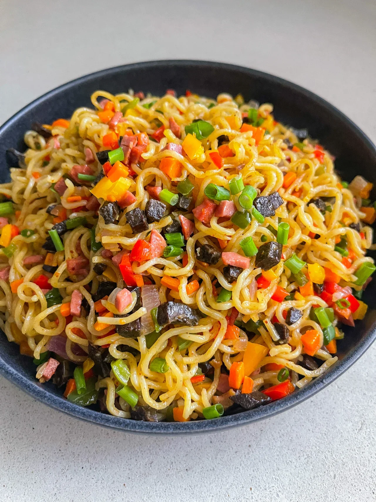 A black bowl filled with cooked ramen noodles mixed with chopped vegetables, diced ham, and green onions.