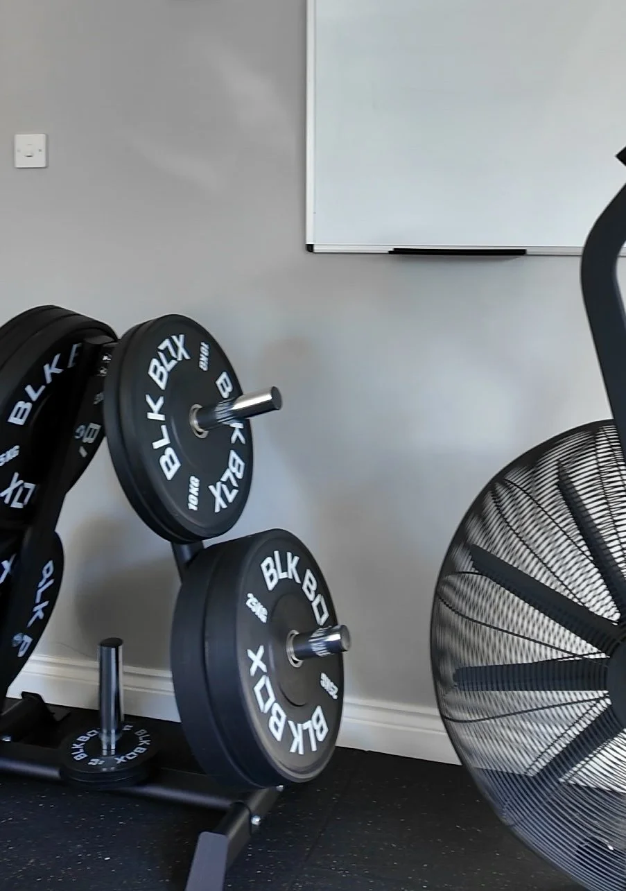 A gym corner with black weight plates labeled 'BLK BOX' on a rack, a large black fan, and a whiteboard on a gray wall.