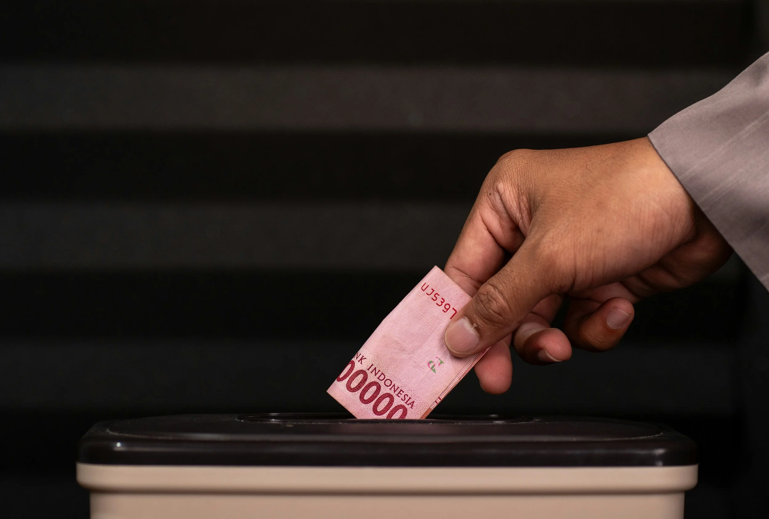 A hand inserting a 100,000 Indonesian Rupiah banknote into a black and white cash deposit machine.