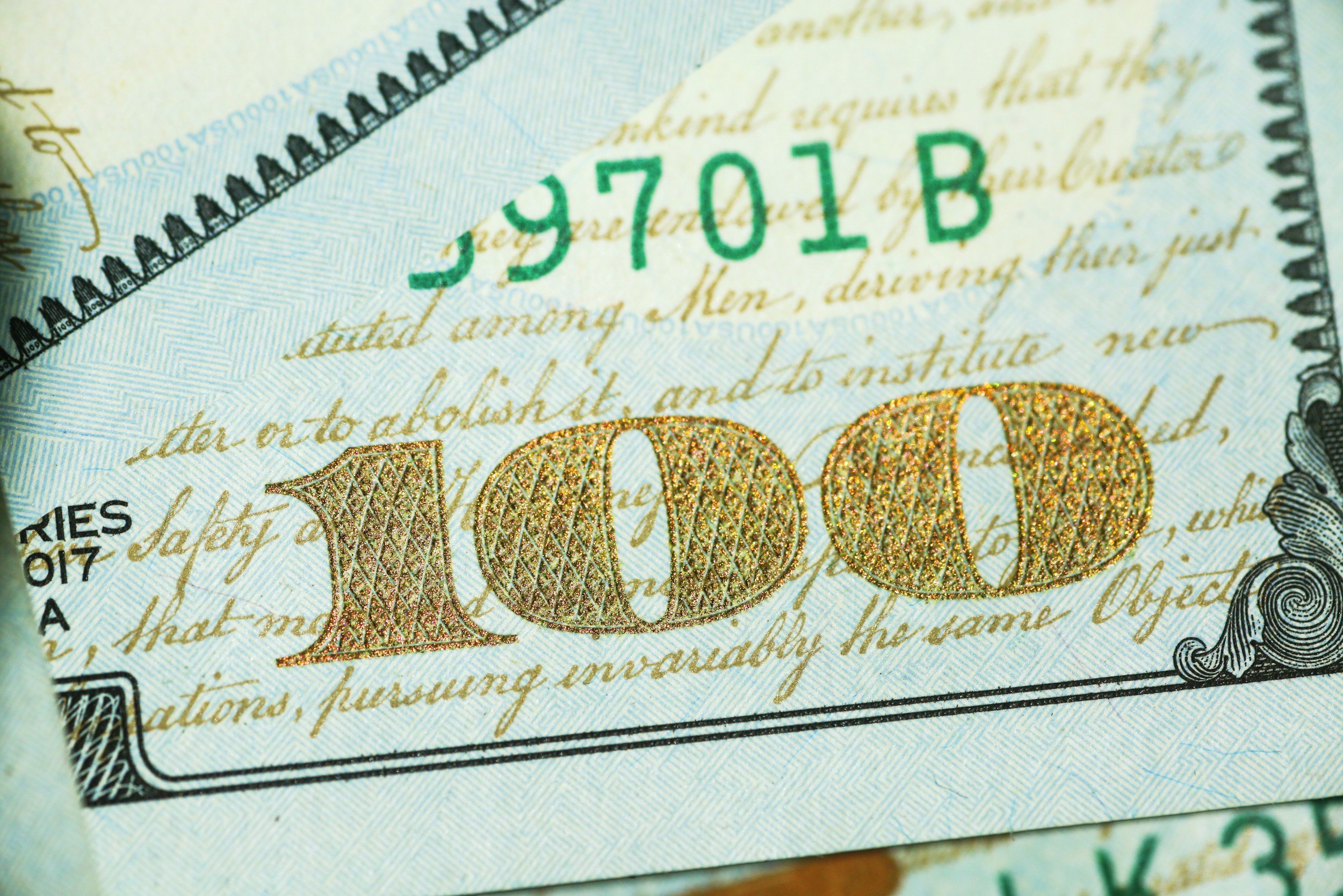 Close-up of a section of a U.S. dollar bill showing the serial number 'S9701B' in green ink, with intricate gold decorative elements and handwritten text in the background.