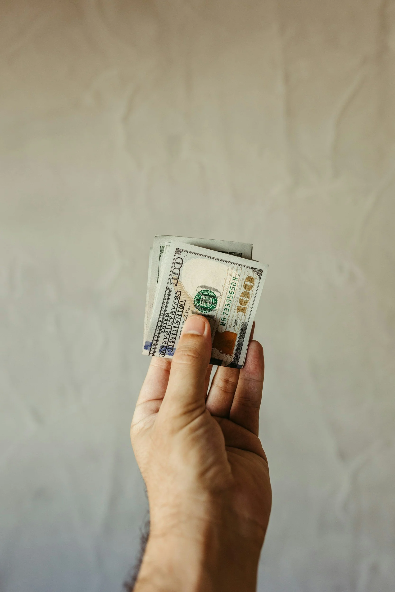 Hand holding a small stack of 100-dollar bills with a plain, light-colored wall in the background.