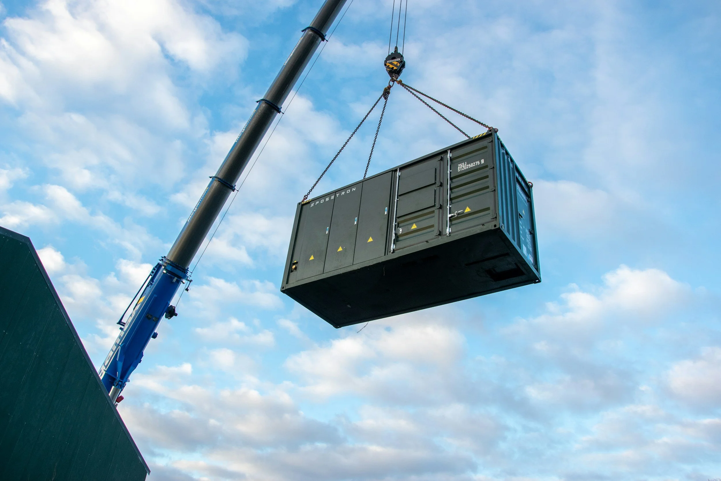A large crane lifting a shipping container in a construction or logistics area outdoors, with a partly cloudy sky in the background.
