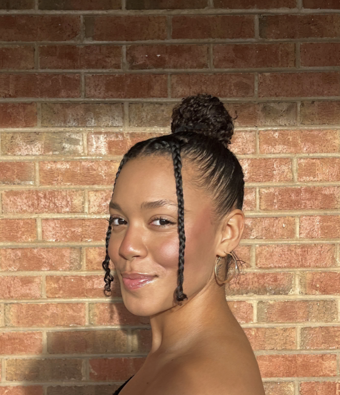 A woman with her hair styled in a high bun and long braids, standing in front of a brick wall.