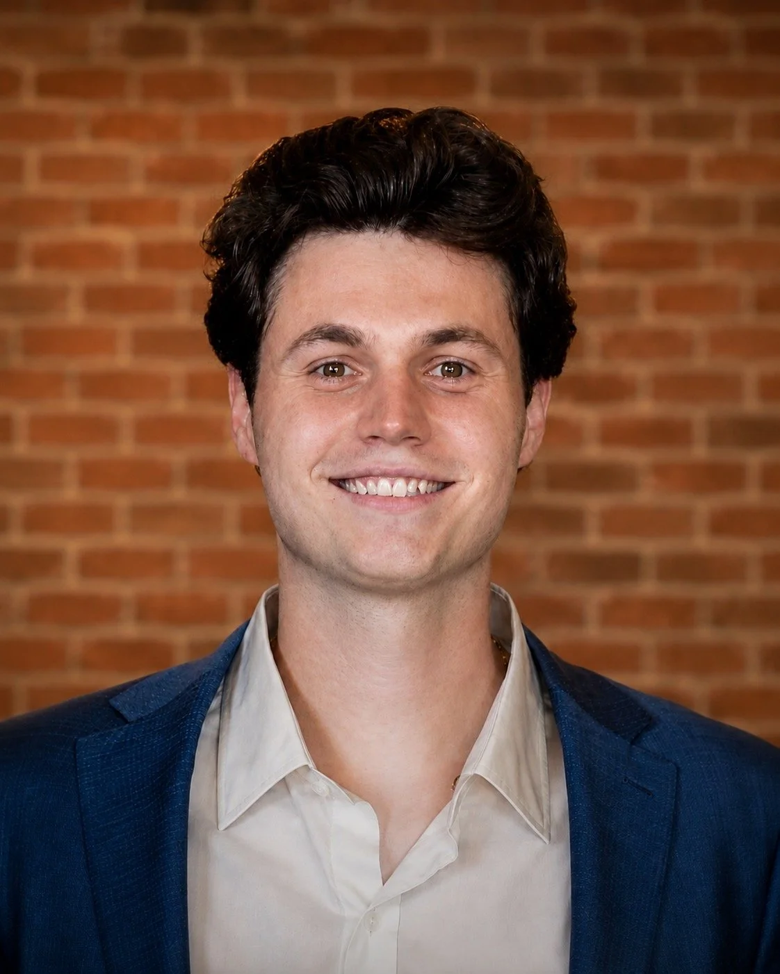 A young man with dark curly hair, wearing a navy blue suit and white collared shirt, smiling in front of a brick wall.