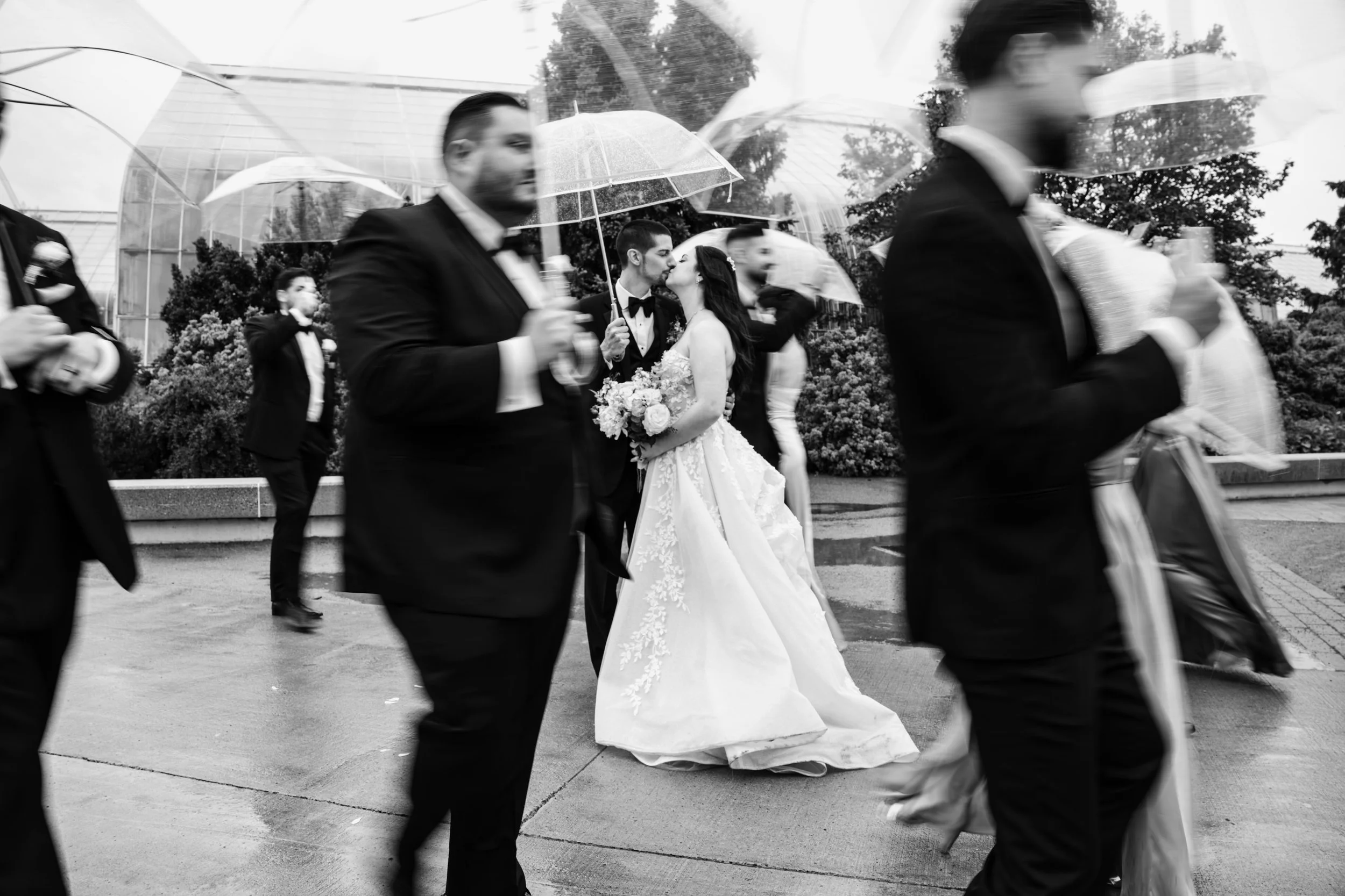 A bride and groom kiss under an umbrella while walking outside on a rainy day, surrounded by wedding guests holding umbrellas.