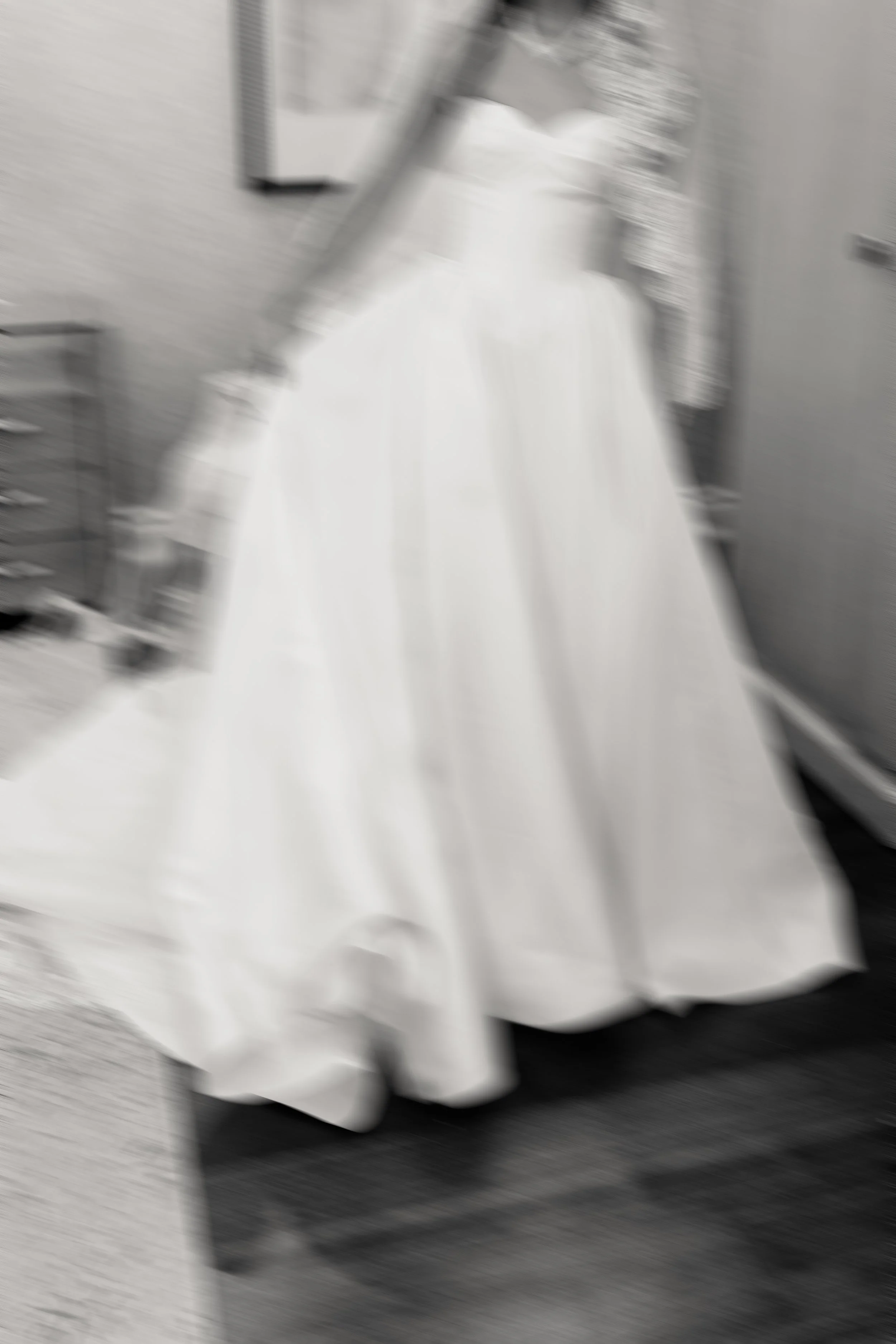 A blurred black and white photo of a dress on a mannequin or hanger, appearing to be a formal gown or wedding dress with a fitted bodice and full skirt.