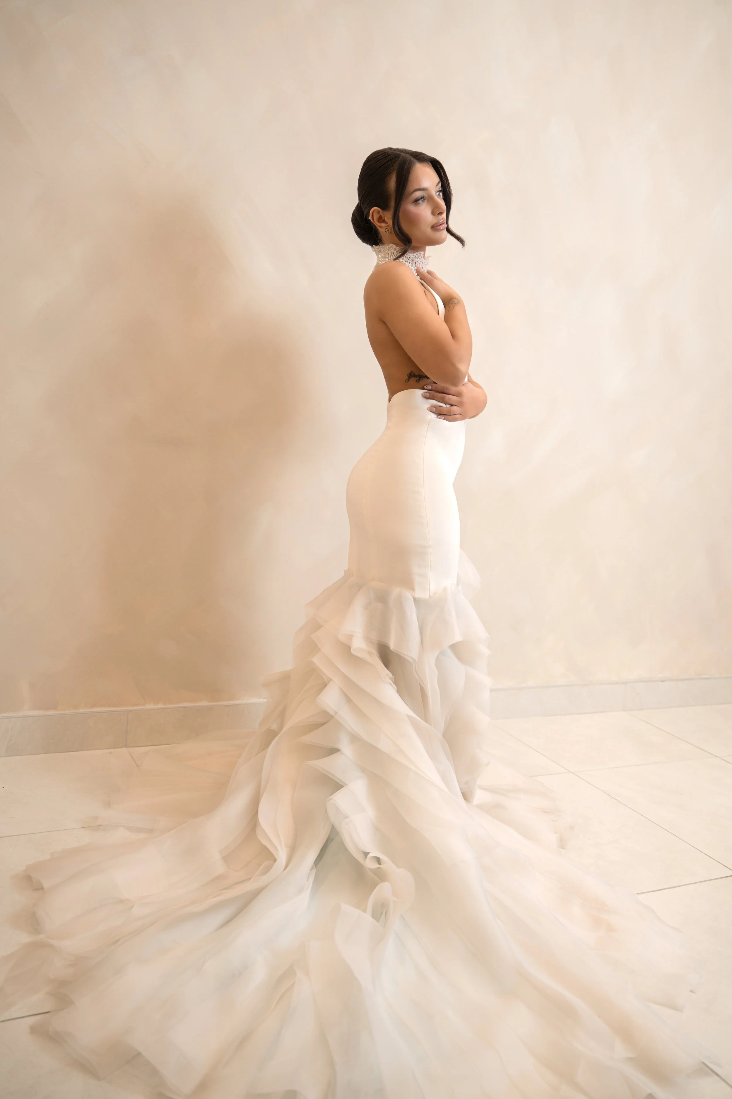 A woman in a white ruffled wedding gown with an off-the-shoulder neckline, standing indoors with a neutral beige wall background, looking to her left.