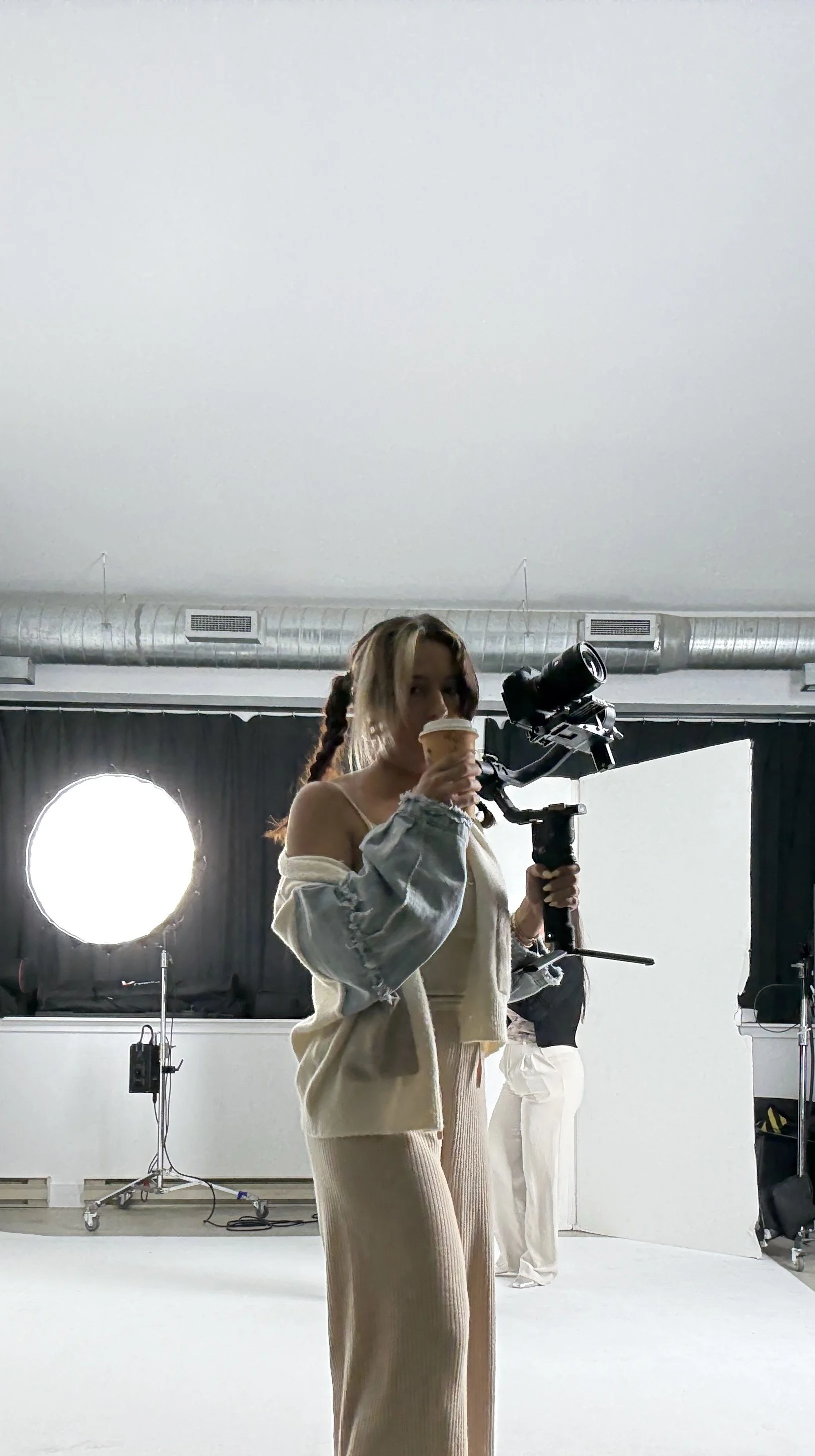 A woman in casual clothing holding a camera in one hand and a cup of coffee in the other, standing in a photography studio with lighting equipment behind her.