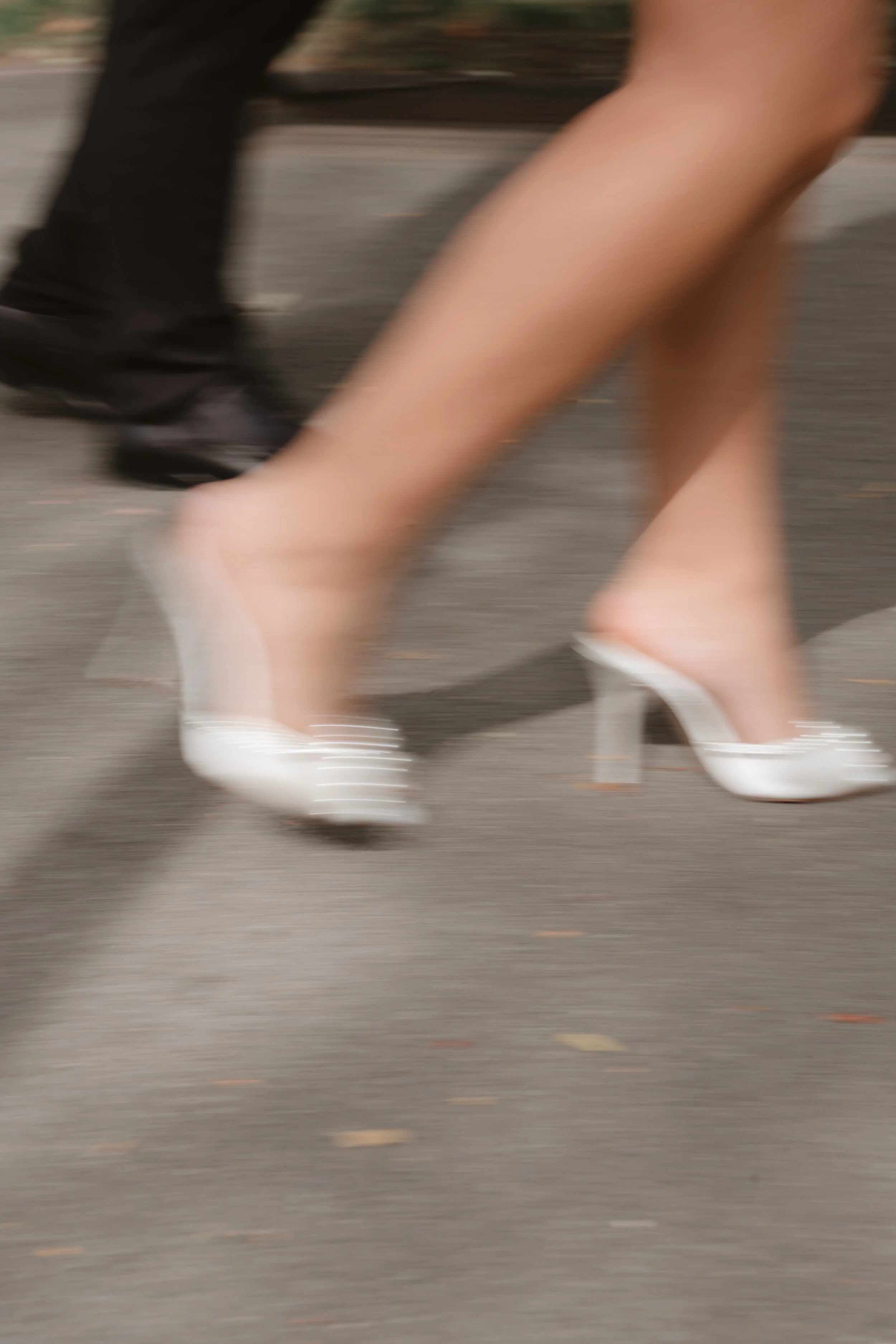 Close-up of a person wearing white high heels walking on pavement, with a blurred background of another person wearing black shoes.