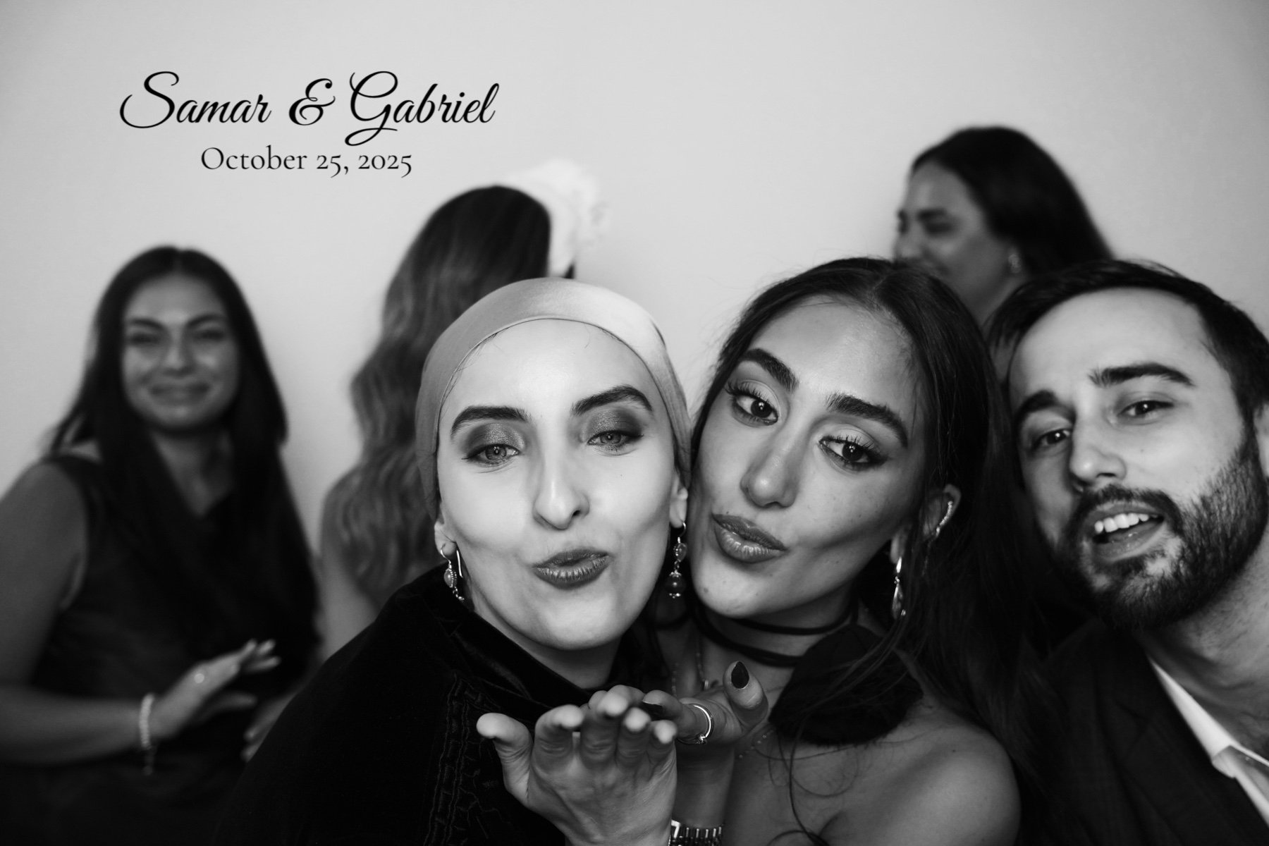Black and white photo of six people at a wedding, with two women posing in the foreground and four others in the background. Text in the top left corner reads "Samar & Gabriel, October 25, 2025."