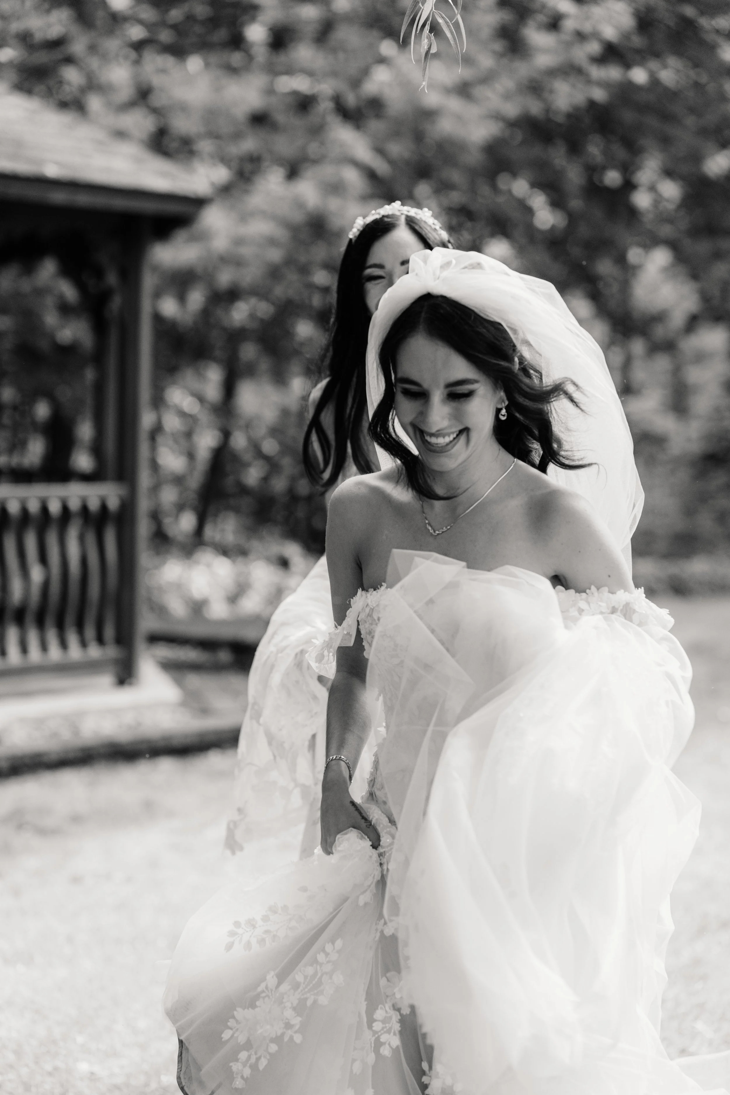 Black and white photo of a bride in a wedding dress with a veil, smiling while holding up her dress, with another woman behind her. The scene is outdoors with trees and a gazebo in the background.
