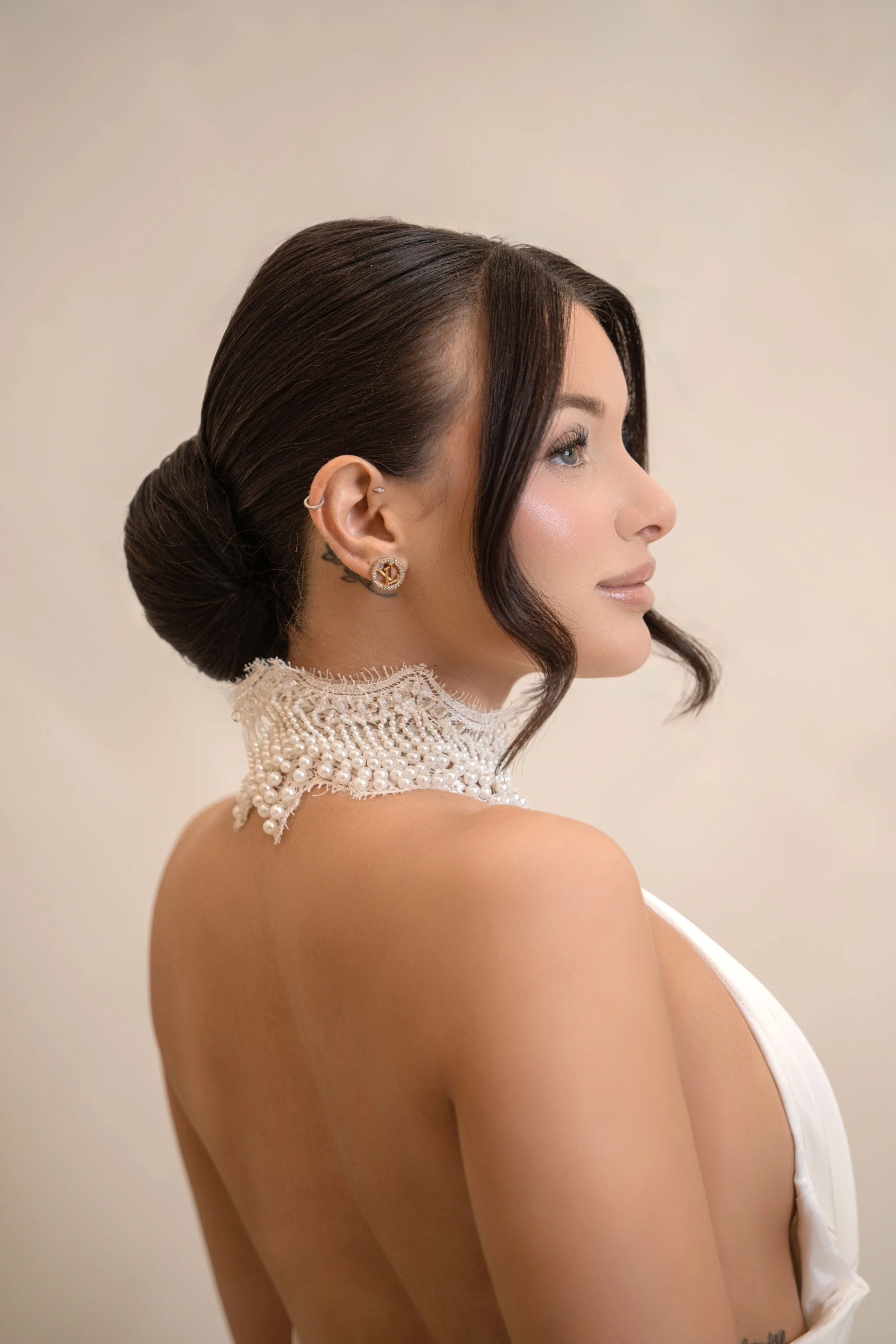 A woman with dark hair styled in an elegant bun, wearing a pearl-adorned lace choker, quick profile view, beige background, and wearing earrings, with makeup highlighting her features.