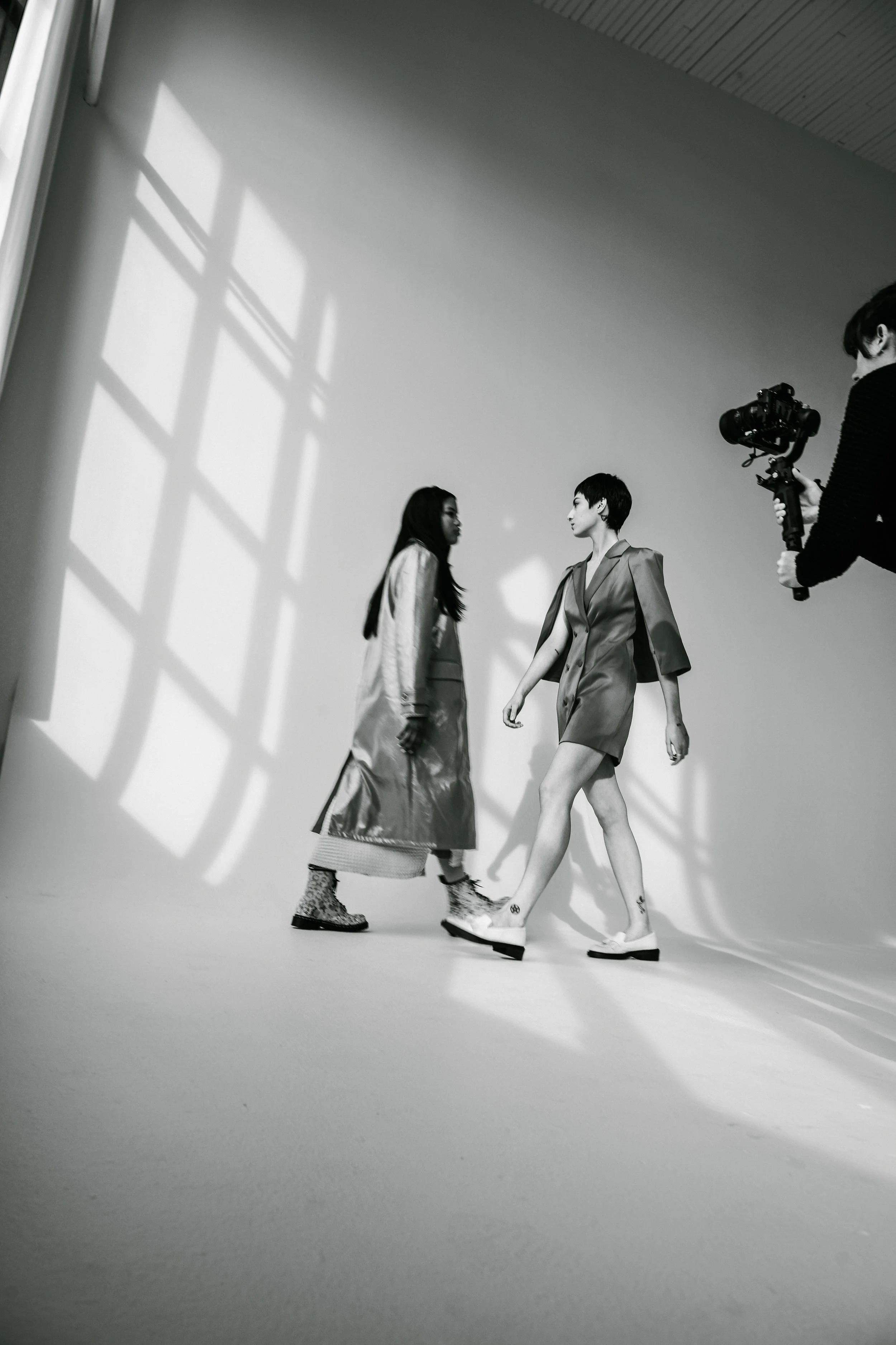 Black and white photo of two women walking in a photo studio, with a photographer capturing the scene. The women are dressed in fashionable clothing, with shadows cast on the wall.