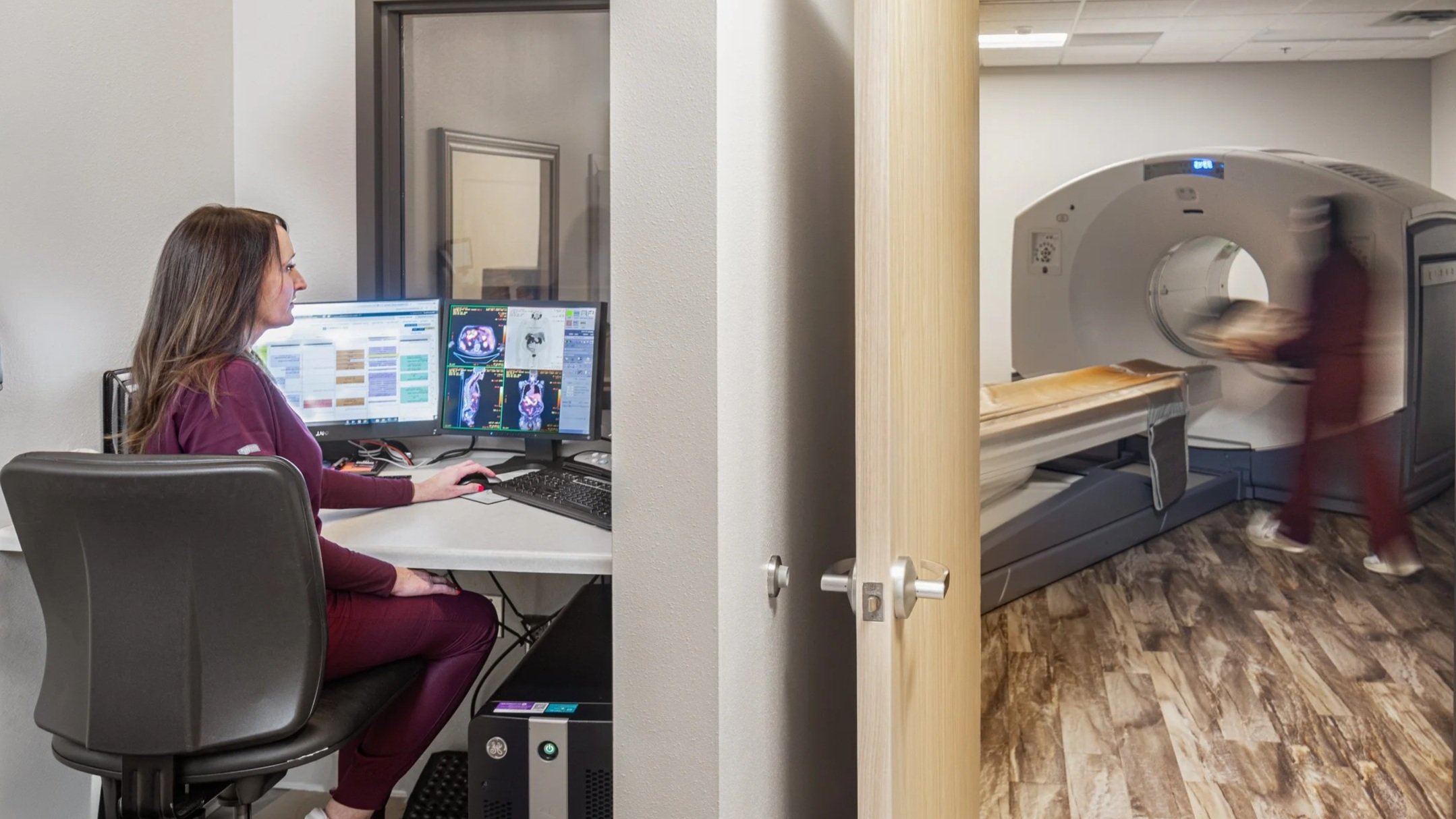 A woman in scrubs sitting at a desk with medical images on her computer screens in a medical imaging room. A patient is walking into a large MRI machine in the adjacent room.