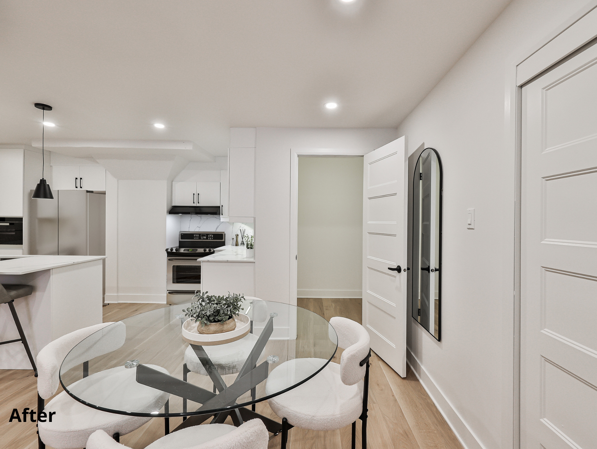 Modern kitchen with white cabinets, stainless steel appliances, and a round glass dining table with white chairs. There is a black pendant light hanging over the island, and a wall mirror next to a white door.