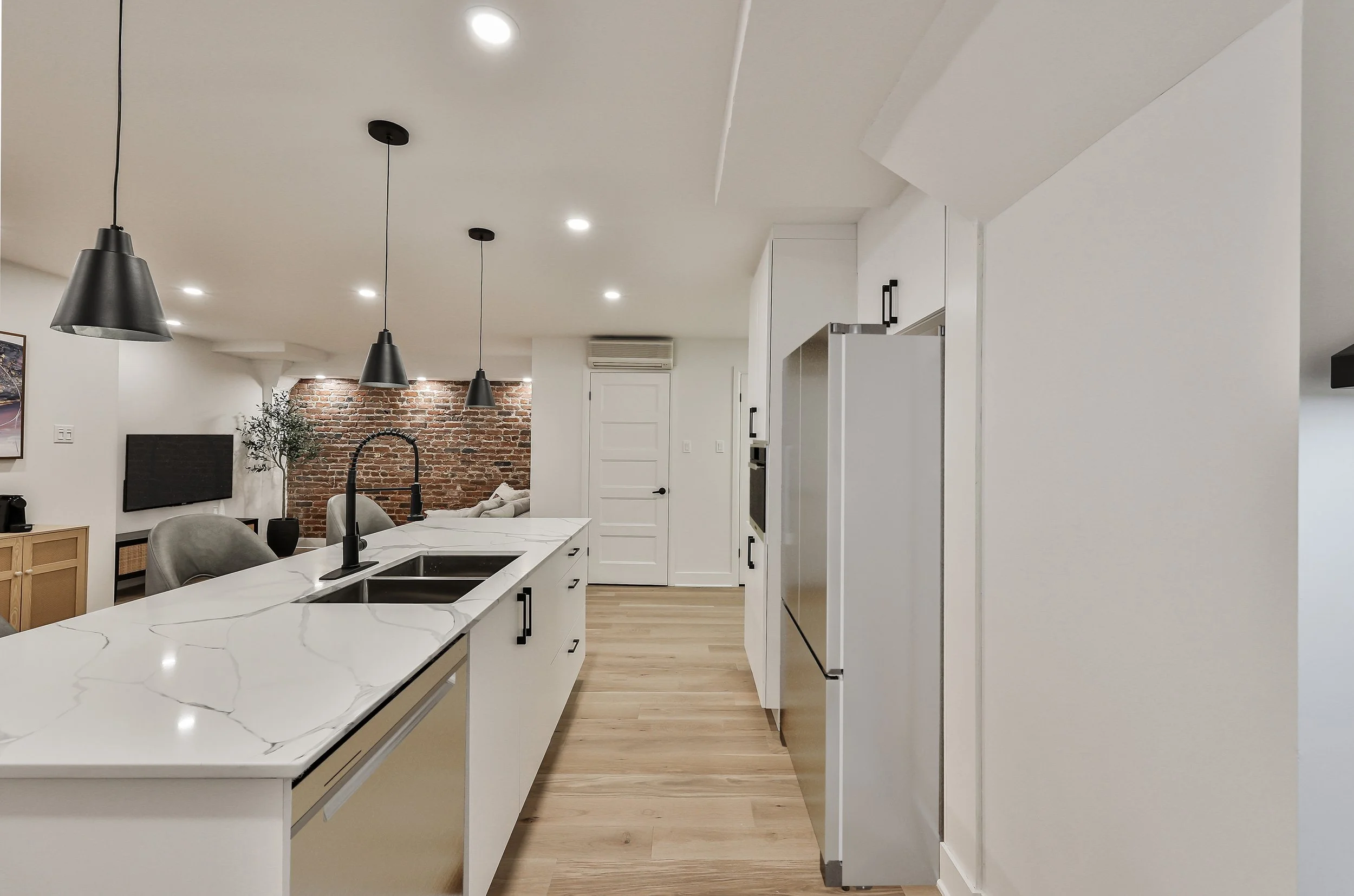 Modern kitchen with white cabinets, stainless steel appliances, and a round glass dining table with white chairs. There is a black pendant light hanging over the island, and a wall mirror next to a white door.
