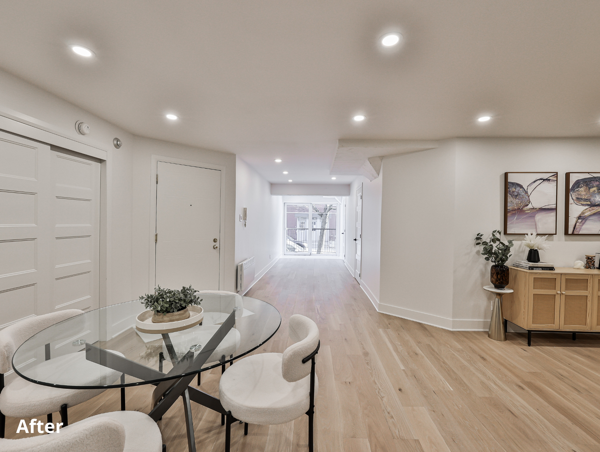 Modern and minimalistic living room with white walls, light wood flooring, and recessed ceiling lights. Features a glass dining table with four upholstered chairs, a wooden sideboard decorated with plants, books, and artwork, and a sliding glass door leading to a balcony.