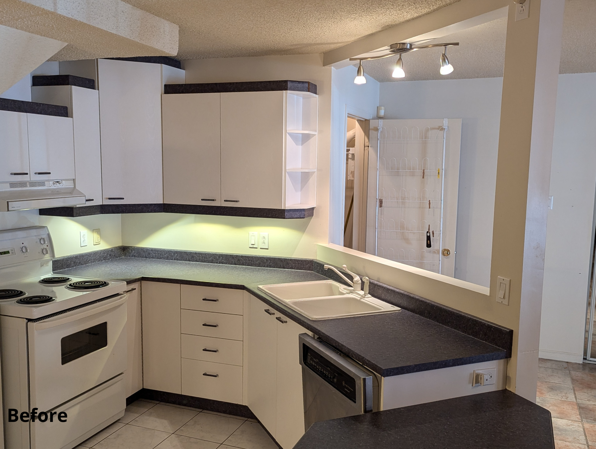 Kitchen with white cabinets, dark countertops, white stove, and a double sink, viewed through a half wall; there are electrical outlets, a silver dishwasher, and a white door with a wire rack in background.