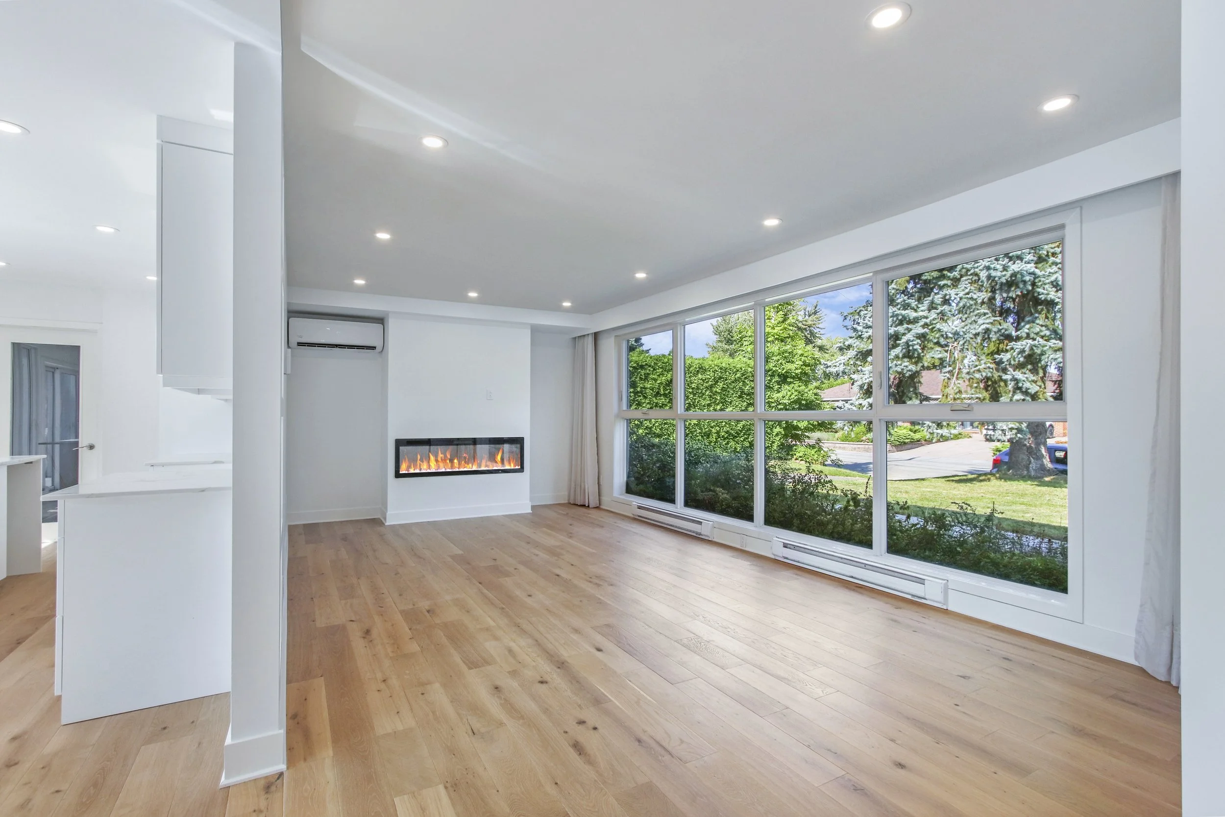 Living room with large windows showing green trees outside, a modern fireplace with flames, white walls, light wood flooring, and recessed ceiling lights.