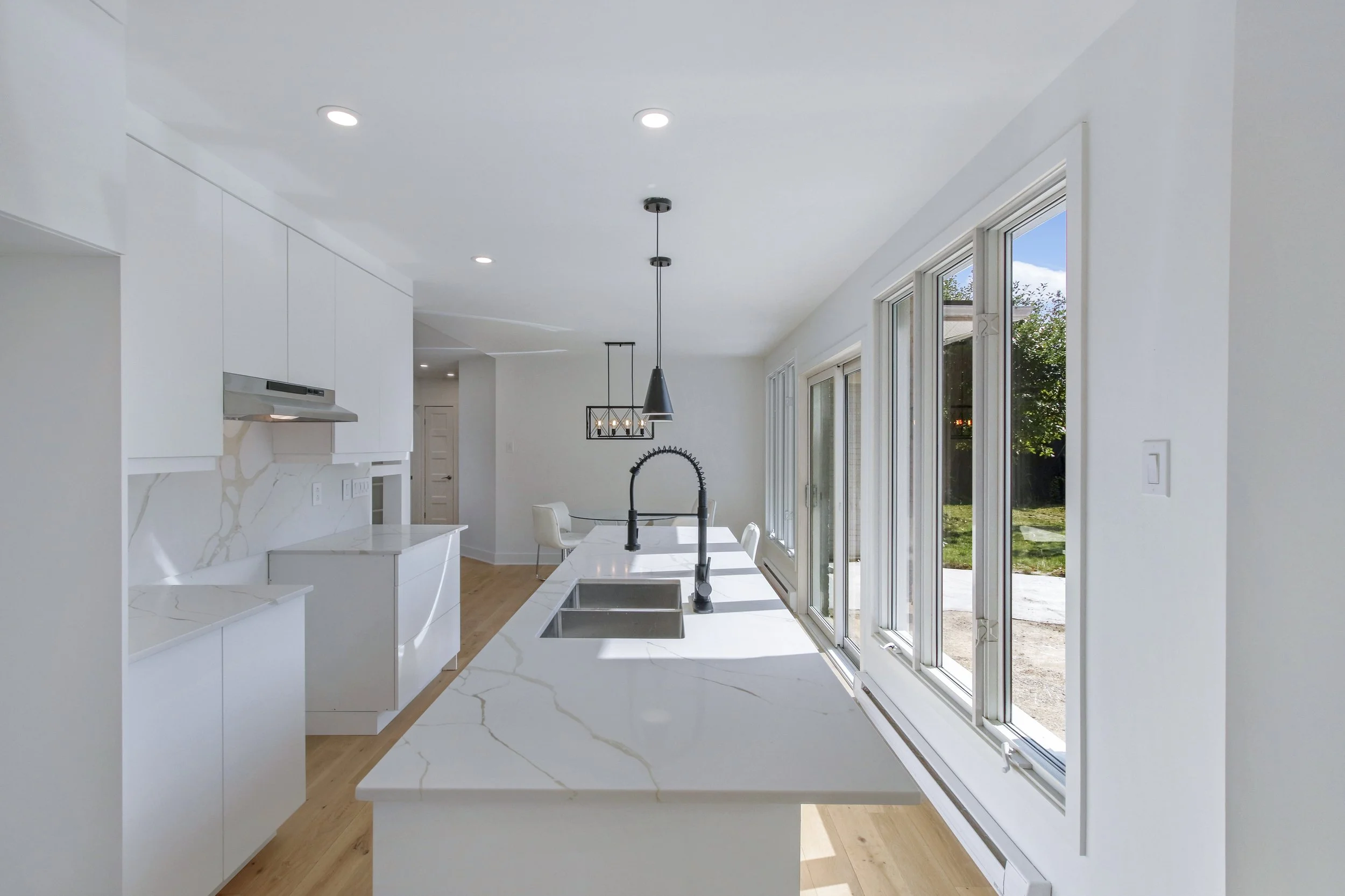Modern kitchen with white cabinets, marble countertops, stainless steel appliances, and large windows overlooking a backyard.