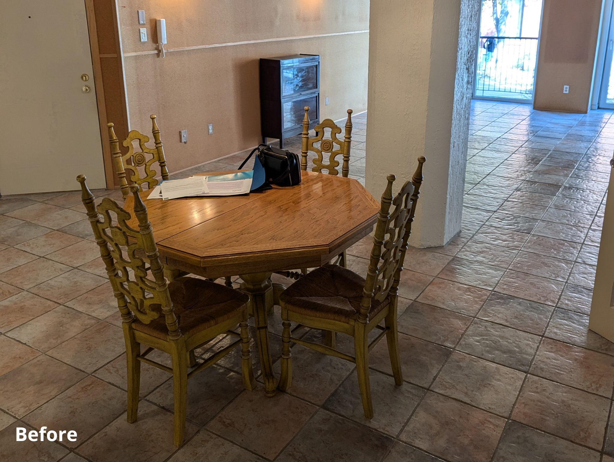 A dining area with a wooden table and four matching chairs with woven seats. Items on the table include a black bag, a book, and some papers. The room has tiled flooring, beige walls, and a small cabinet against the wall. There is a door on the left and a hallway leading to a bright area with a sliding glass door and balcony outside.