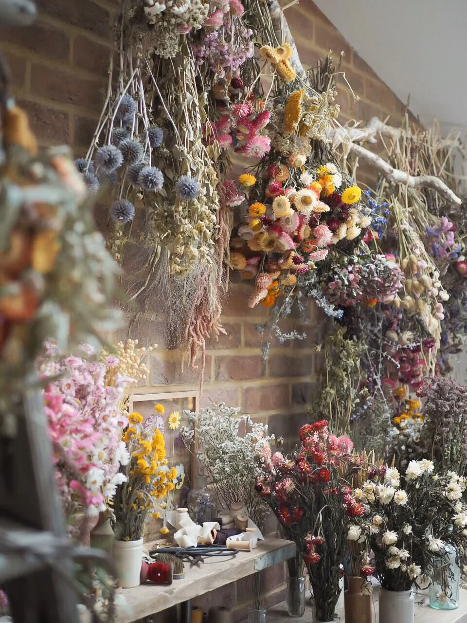 Wall adorned with hanging and standing dried flowers in various colors, including pink, yellow, white, purple, and red, on a rustic brick wall inside a florist's workshop.