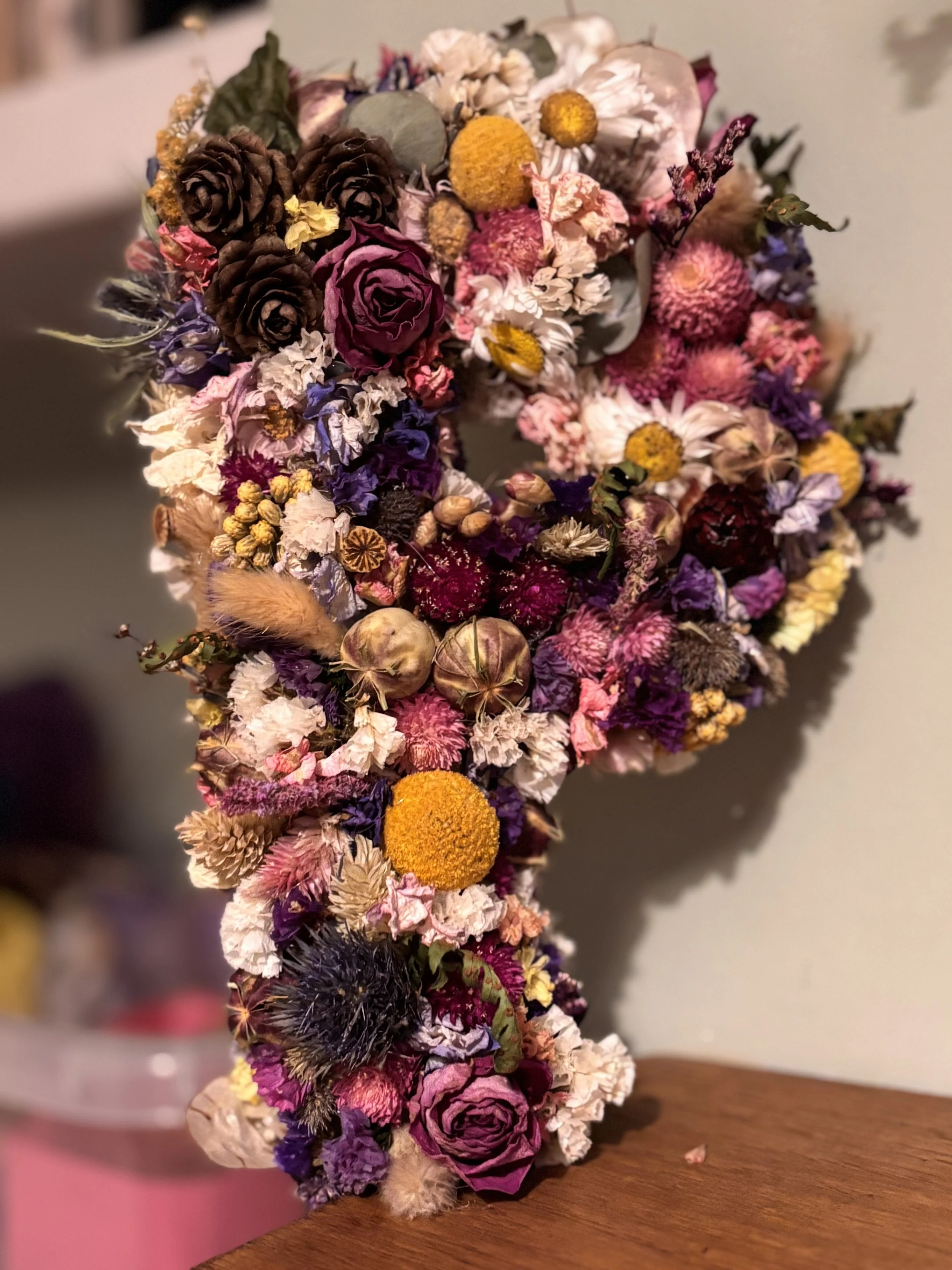 Colorful floral arrangement shaped like a letter 'J' with dried and fresh flowers, including roses, daisies, and pinecones, on a wooden surface.