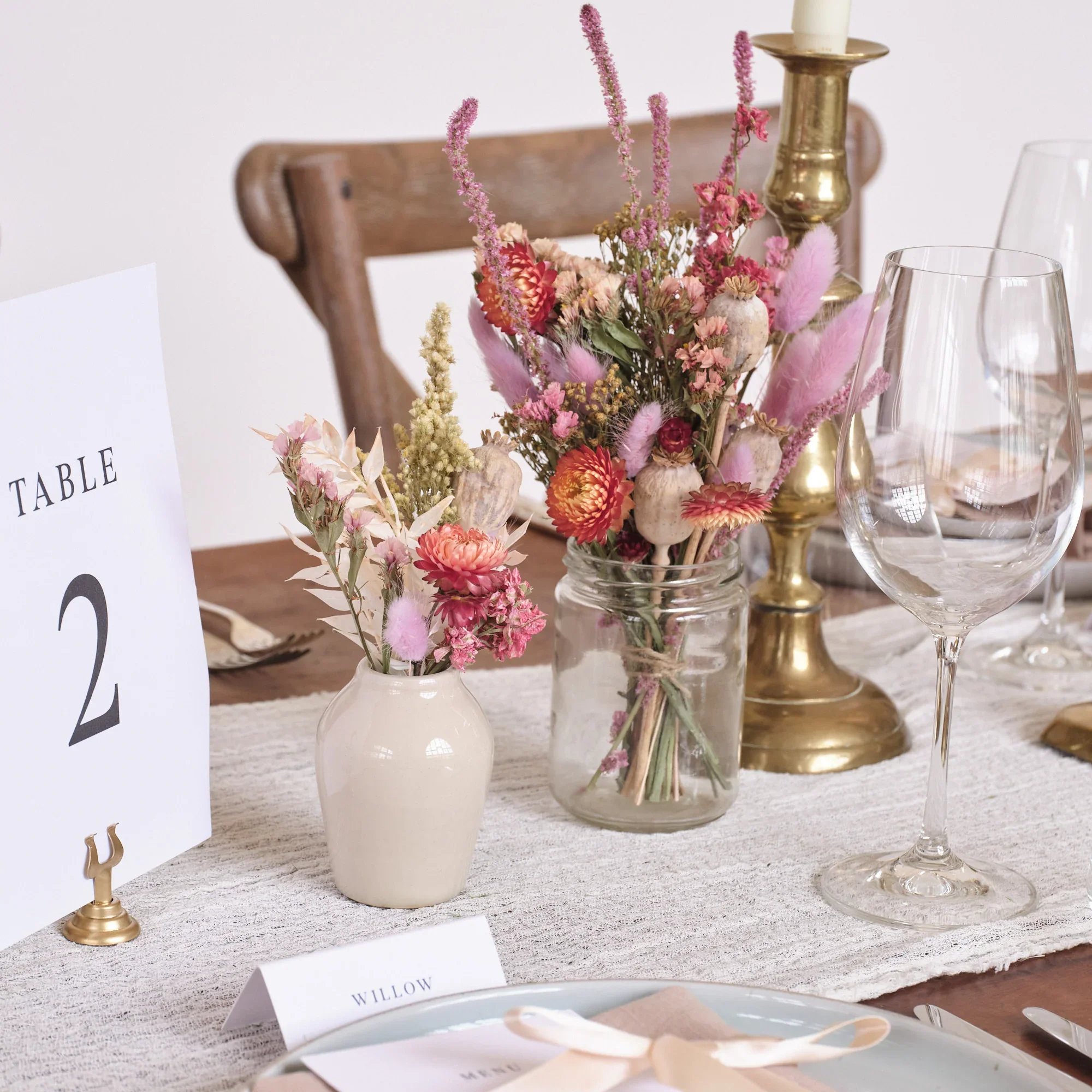 A floral centerpiece with pink, red, and purple flowers in a jar, set on a dining table with a white table runner, a white place card labeled 'Willow,' and empty wine glasses, decorated with a brass candlestick and small decorative items.