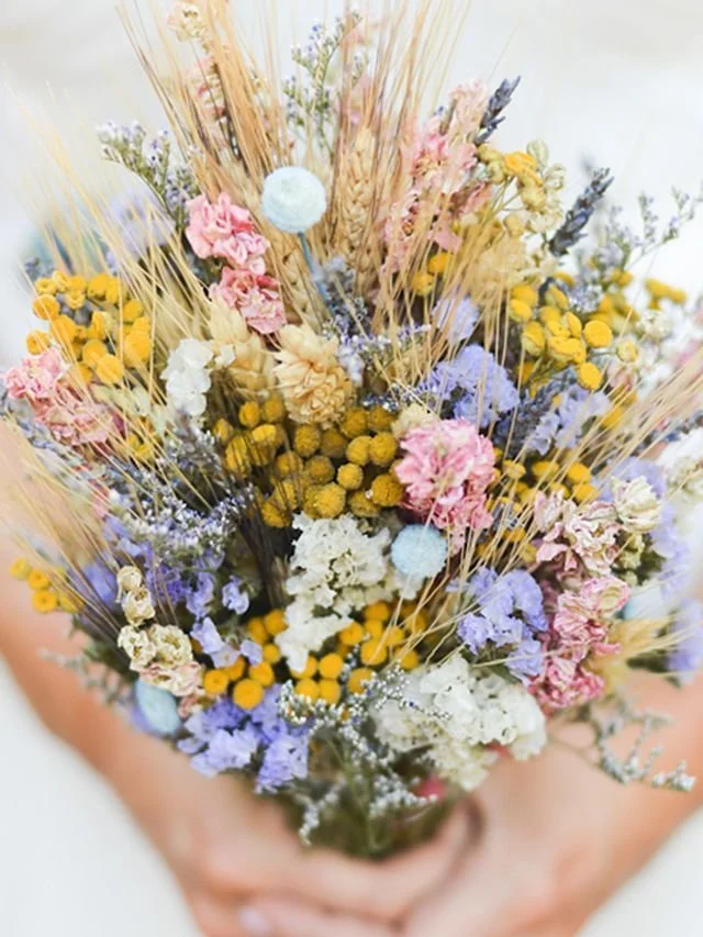 A bouquet of dried flowers with pink, yellow, white, purple, and blue blossoms, and dried grasses being held by a person.
