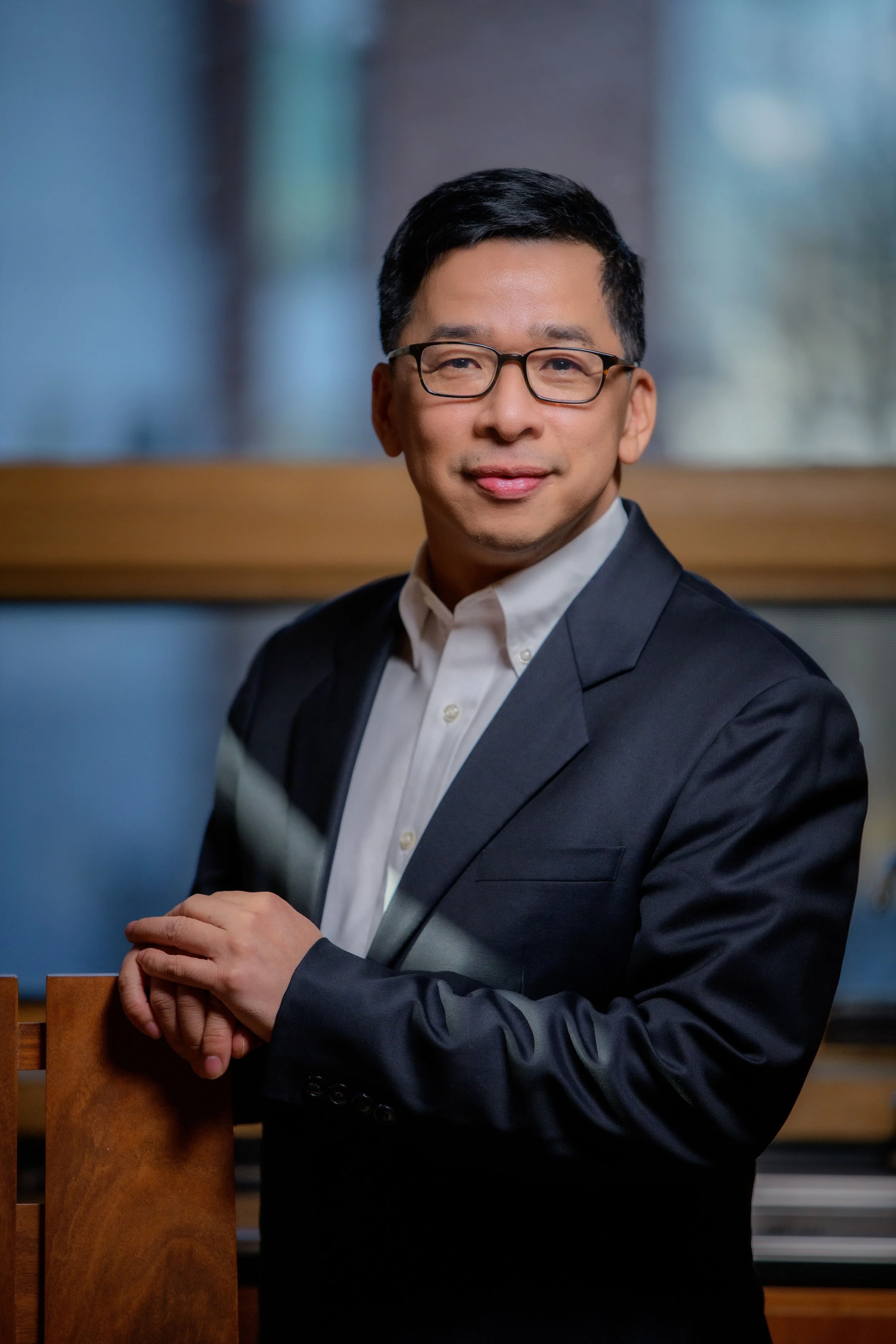 A man wearing glasses, a dark suit, and a white shirt, standing in a professional setting with a blurred background, looking at the camera, smiling slightly, with hands resting on a wooden surface.