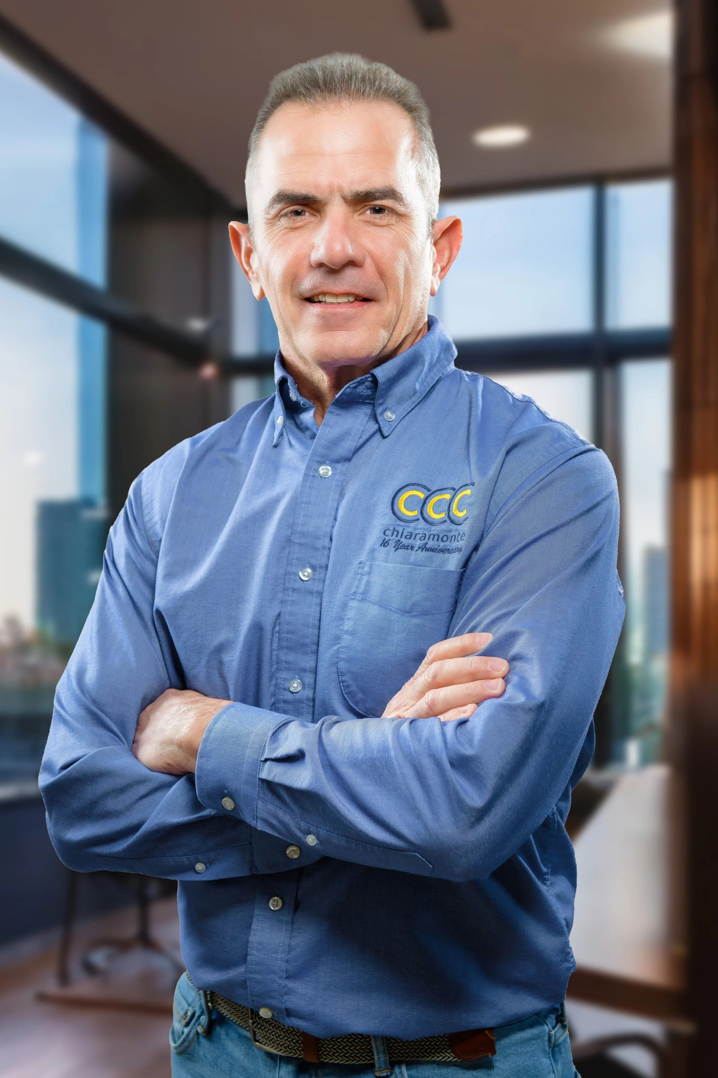 A man with gray hair, dressed in a blue button-up shirt with a logo on it, standing with arms crossed in an office setting with large windows and natural light.