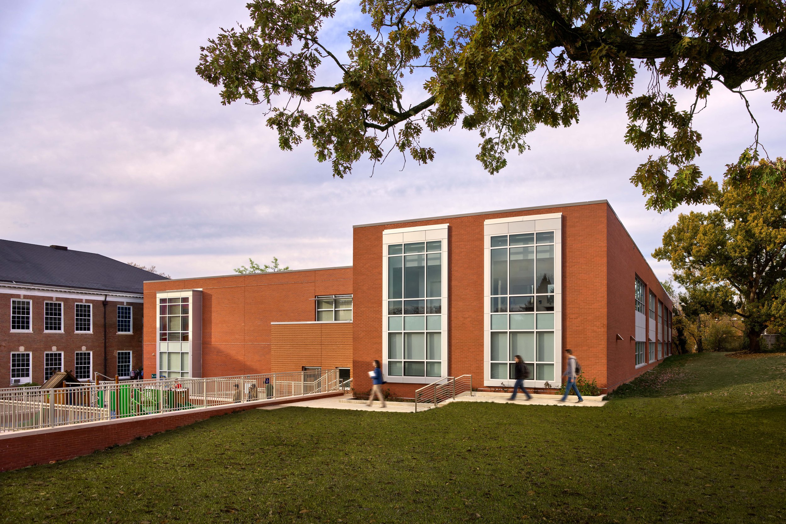 A modern red brick school building with large window panels, surrounded by green trees and a grassy yard where students are walking.