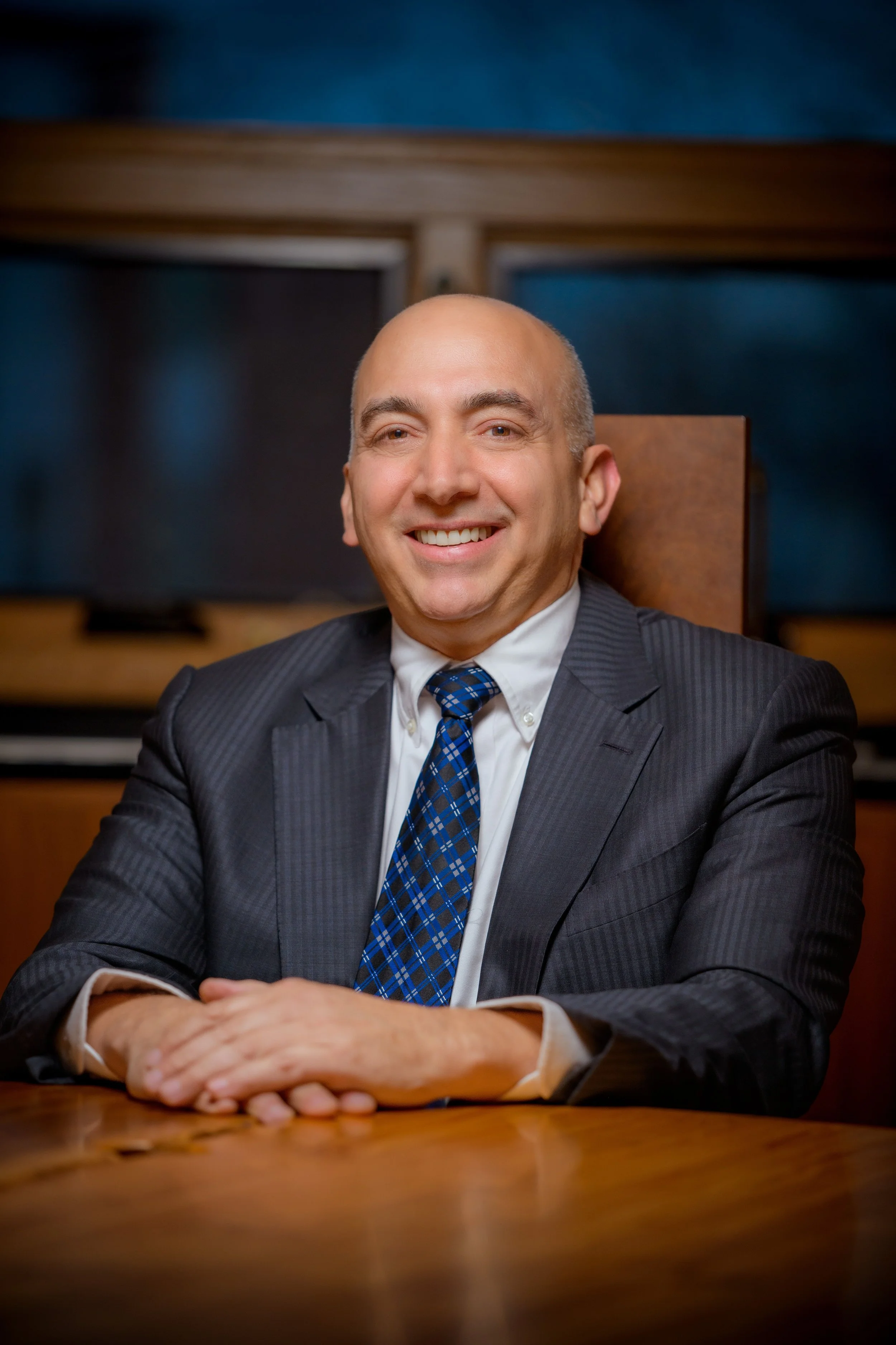 A smiling man in a suit and tie sitting at a wooden desk with his hands folded, in front of a background with blue walls and a wooden conference table.