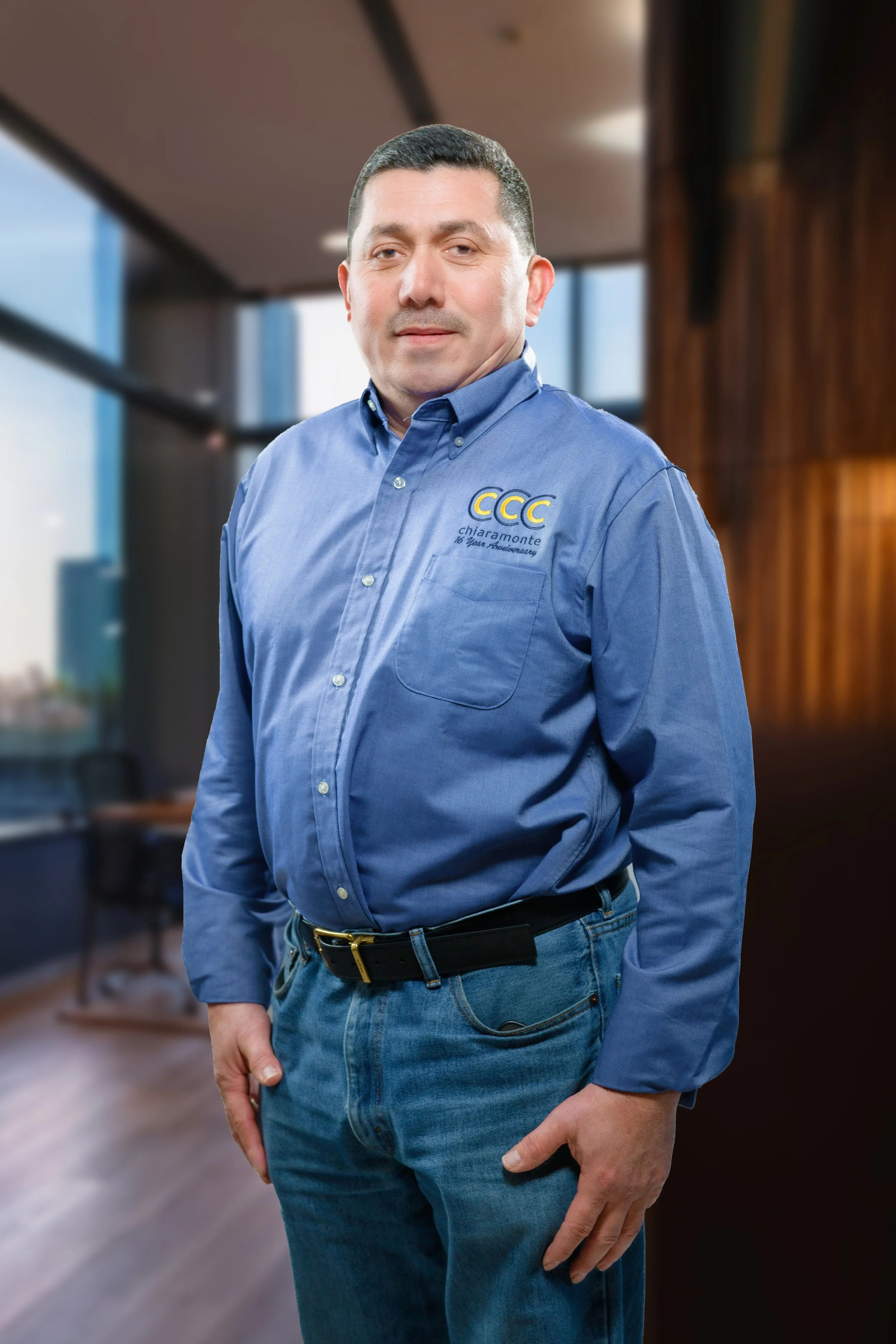 A man with short dark hair and light skin wearing a blue button-up shirt with a logo and text, standing in a modern office space with large windows and wooden wall panels.