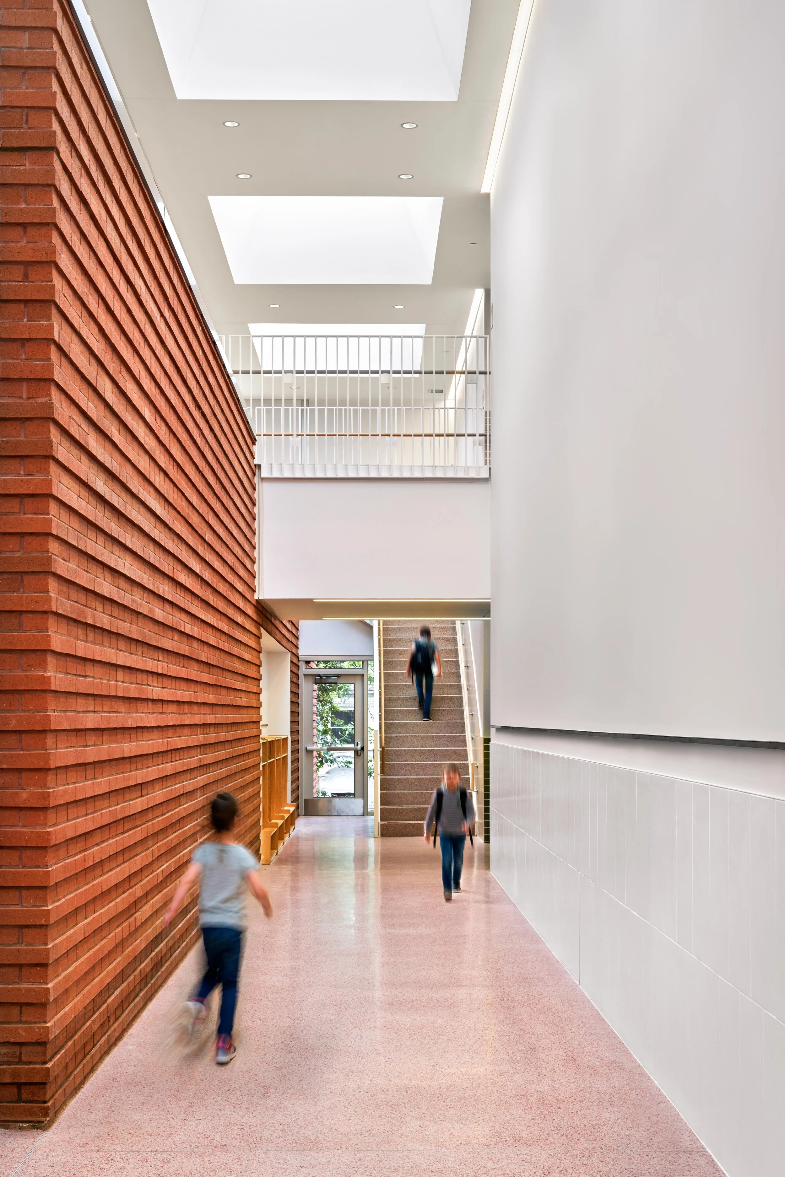 Interior of a modern building with a red brick wall on the left, white walls on the right, a staircase in the middle, and several people walking through the hallway.