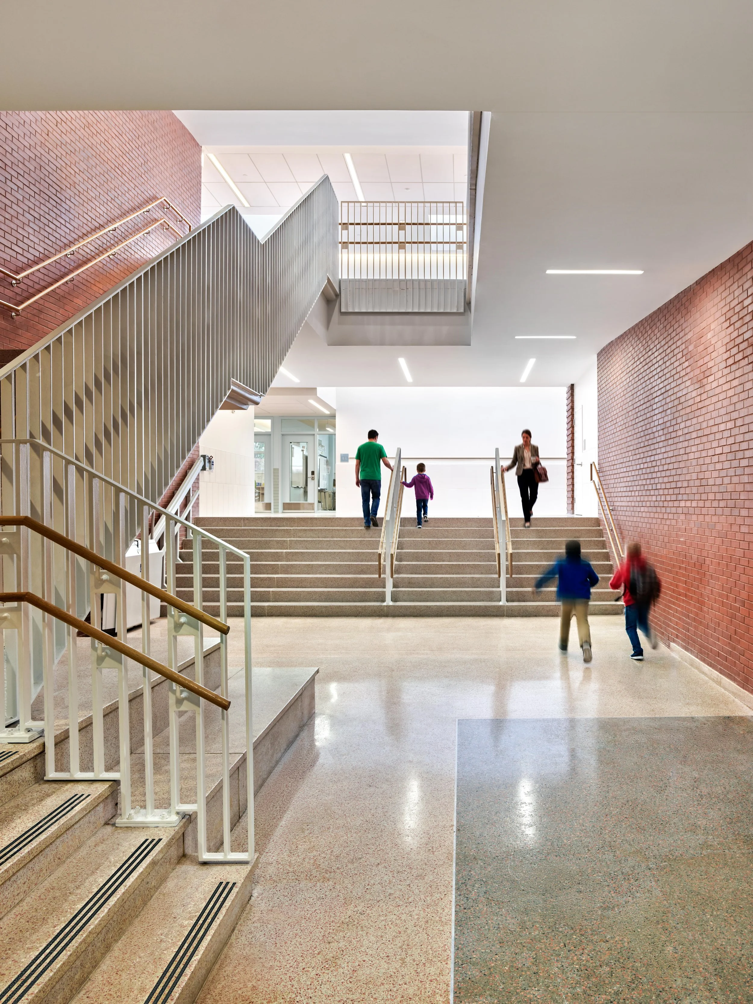 An indoor staircase in a modern building with a brick wall on the right and a white wall on the left. Several people, including children and adults, are walking up and down the stairs.