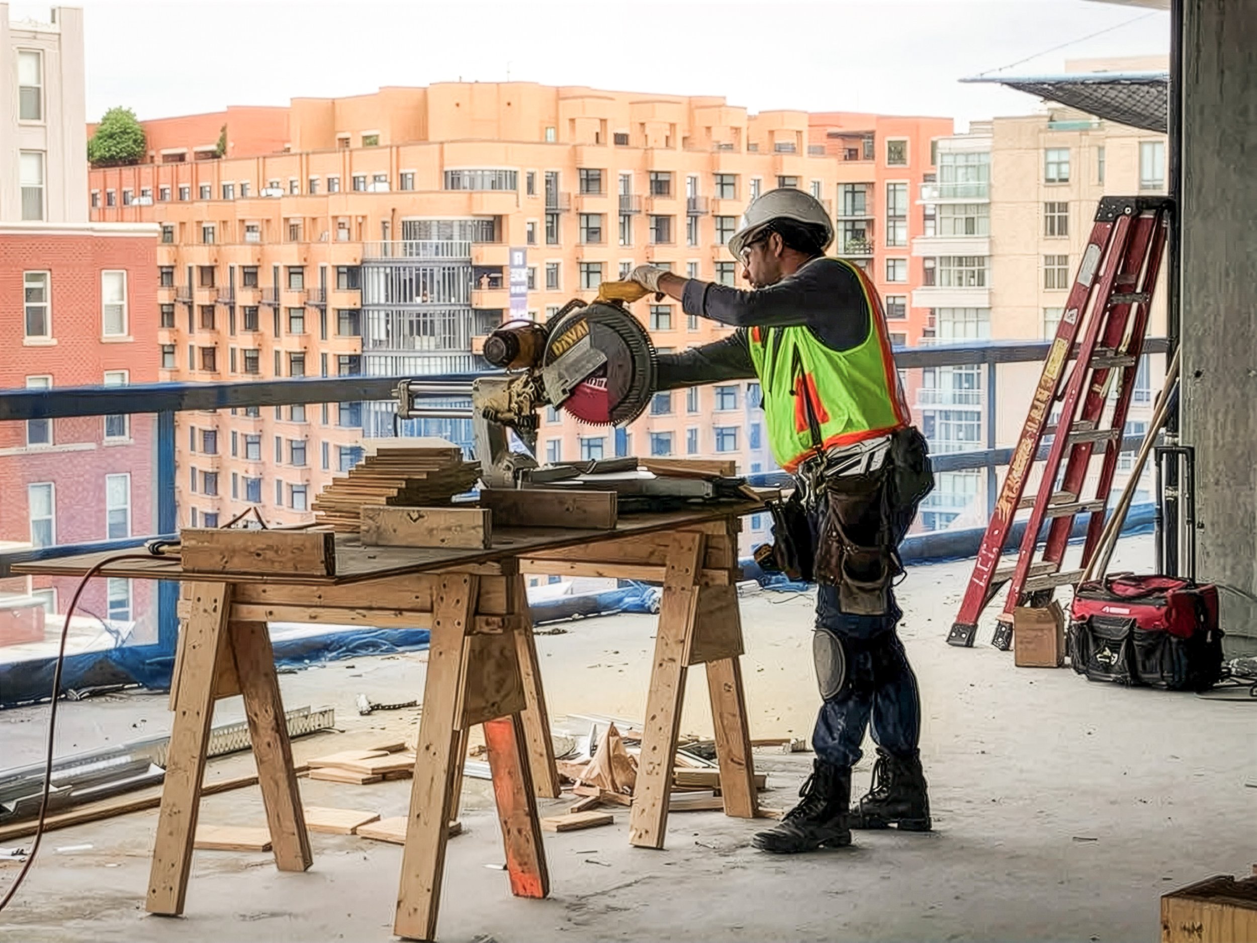 Construction worker using a power saw on a worktable at a building site, with residential buildings in the background.