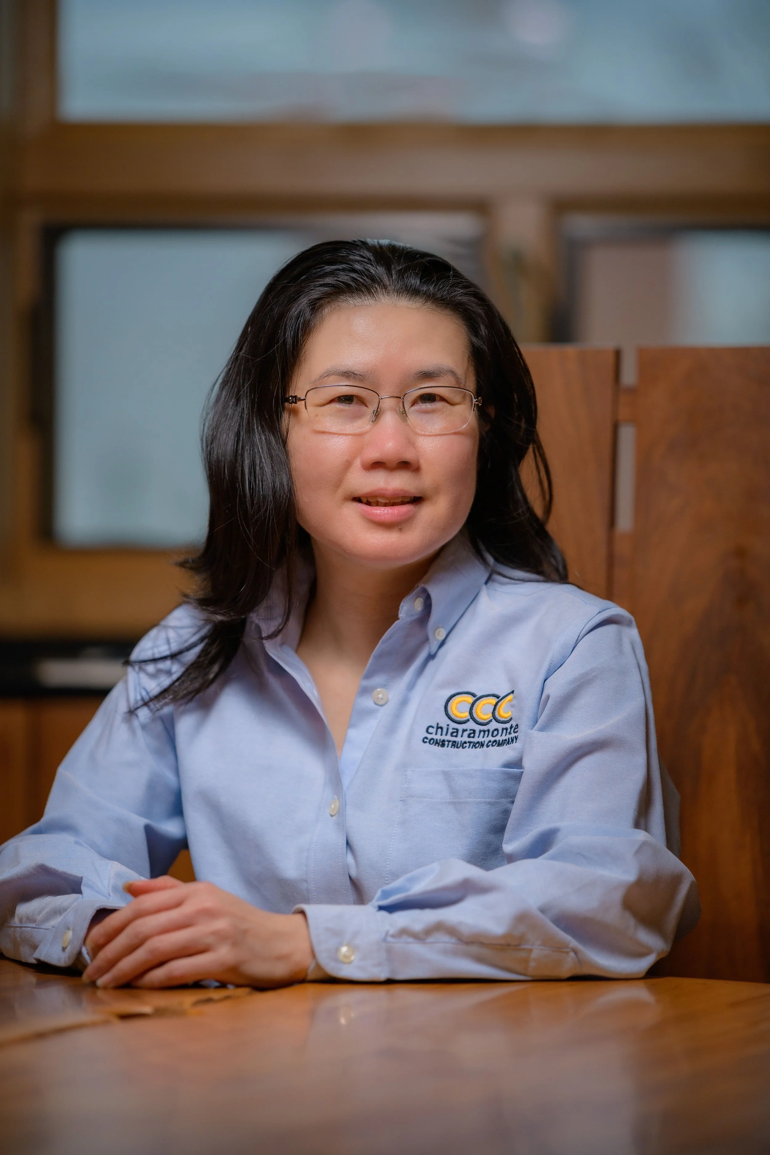A woman with shoulder-length black hair, glasses, and a light blue button-up shirt featuring the logo of Chiaramonte Construction Company, sitting at a wooden table in a room with wood-paneled walls and a window.