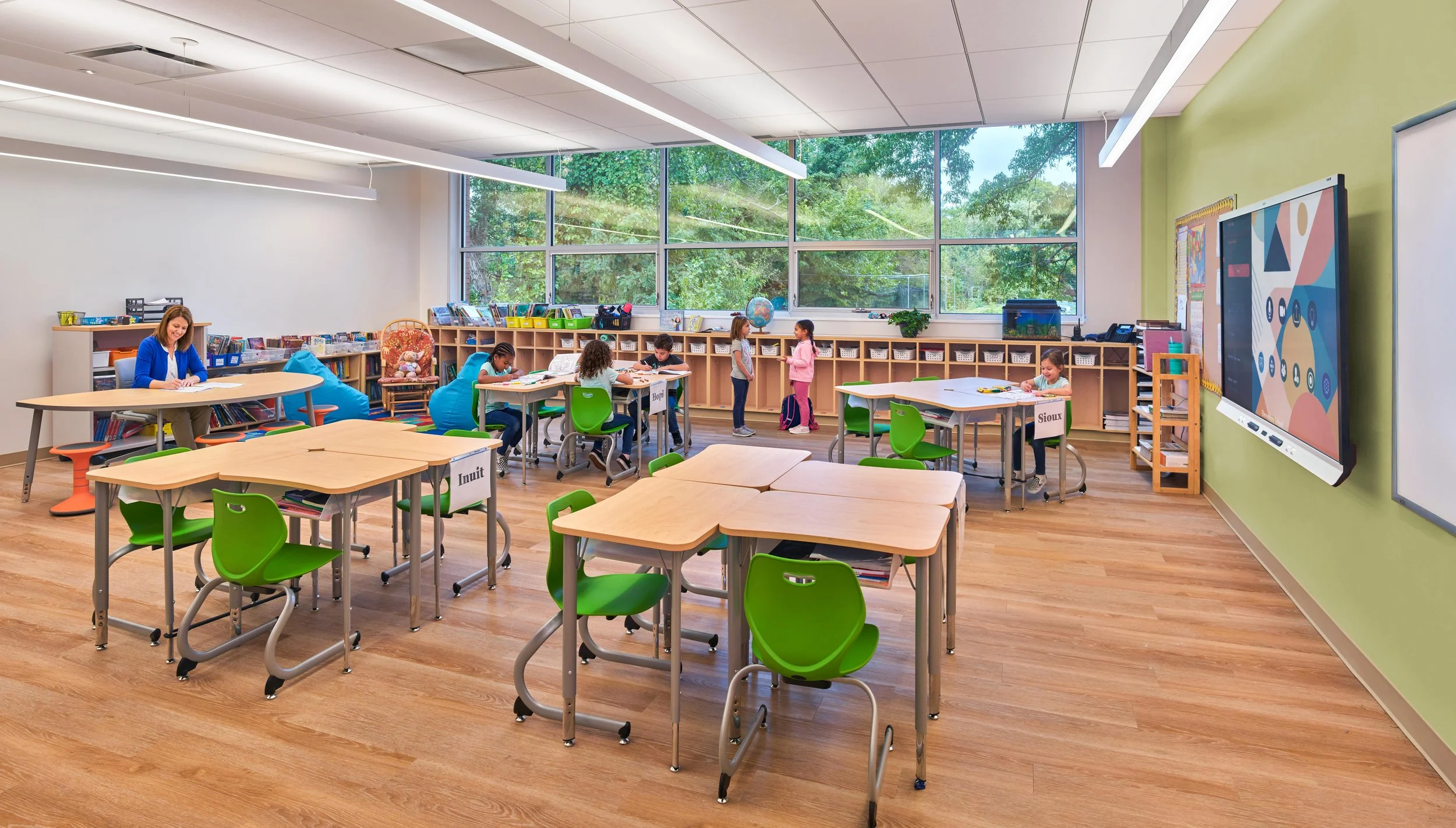 A bright, spacious classroom with large windows overlooking trees. Inside, children sit at small green desks, working or standing. A teacher is seated at a table near bookshelves, with educational materials and colorful decorations around.