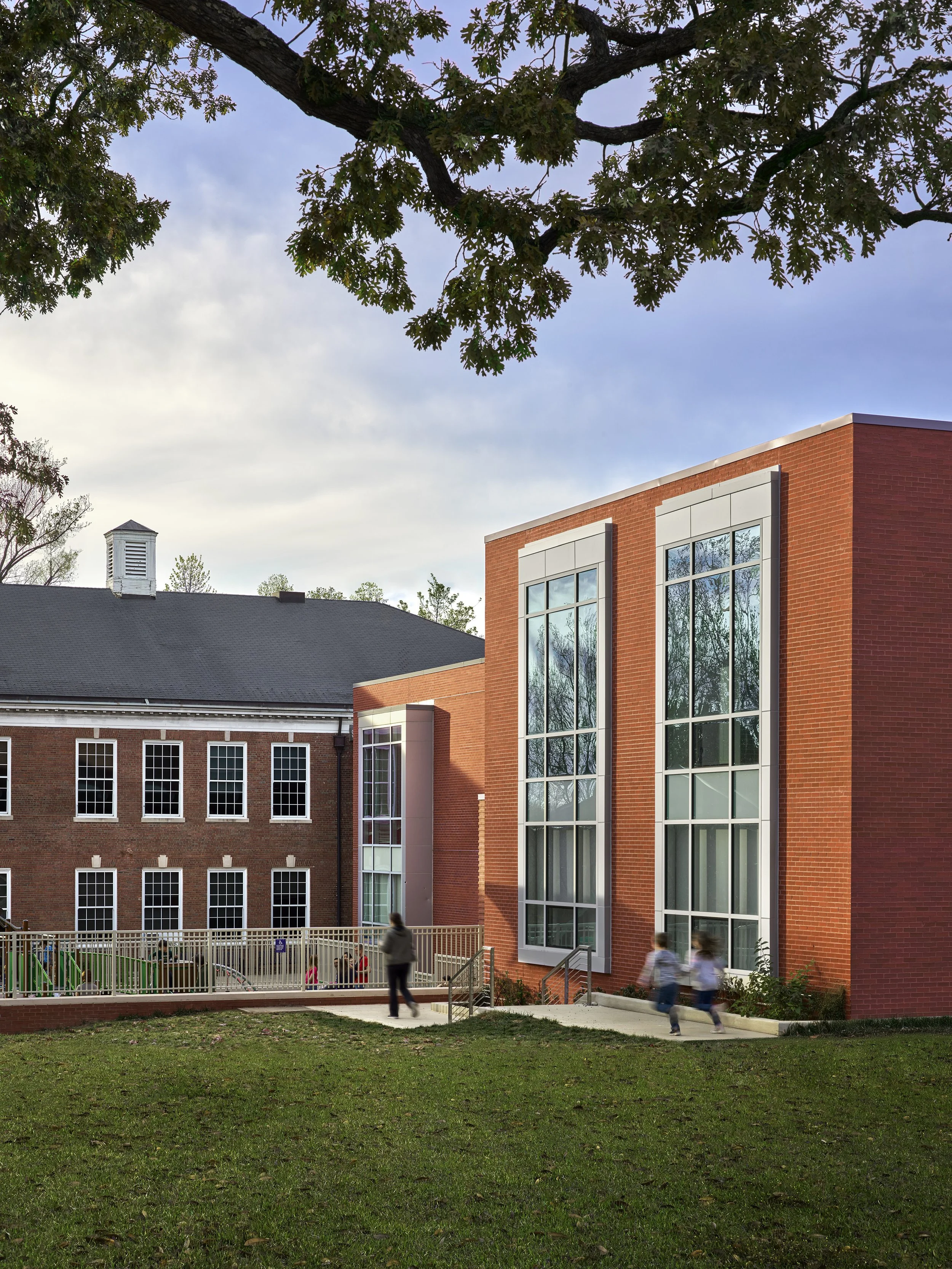 A school or university building made of red brick with large glass windows, surrounded by green lawn and trees, with children and a few adults walking nearby on a sidewalk.
